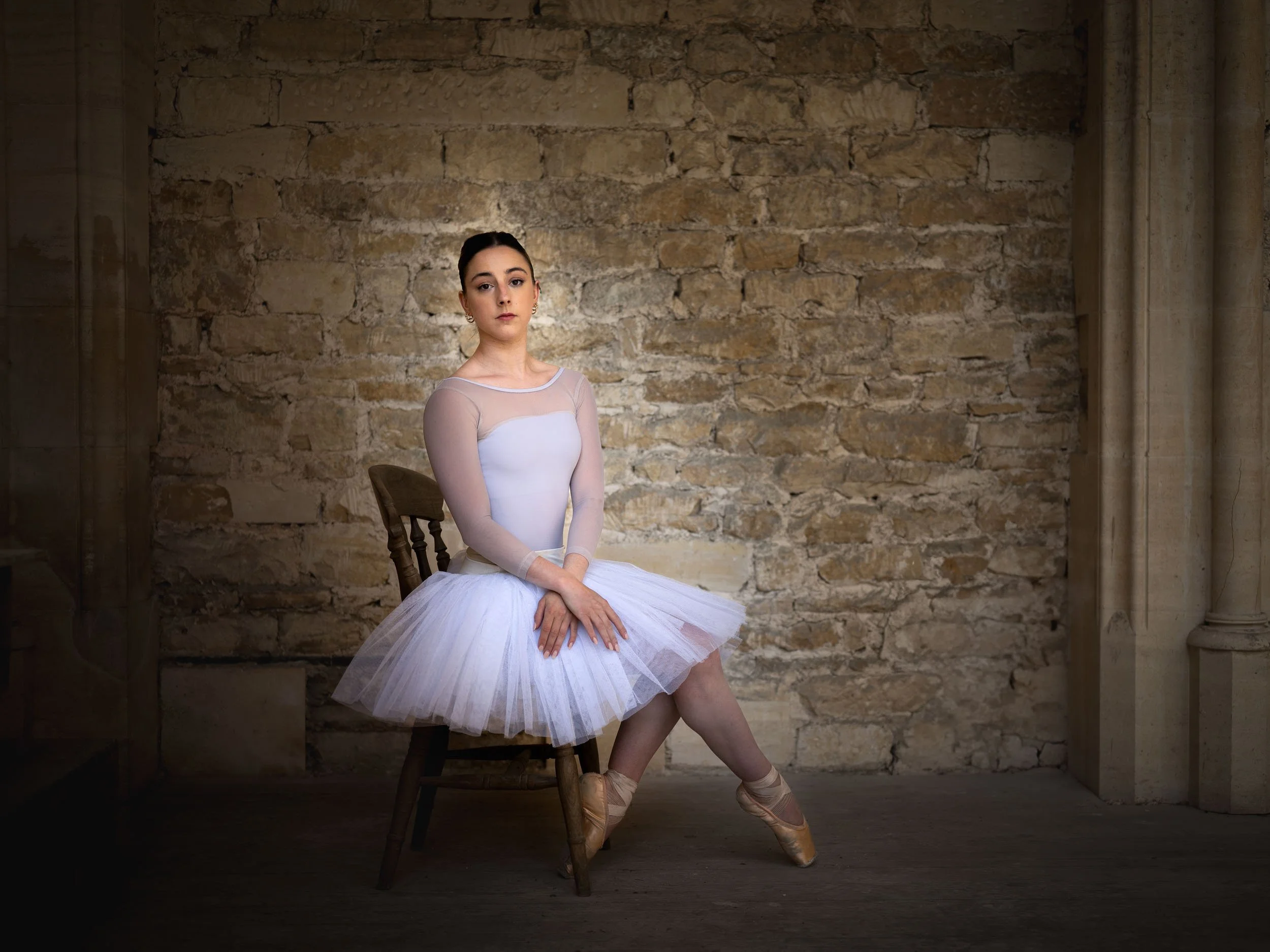 Ballet dancer sitting on a wooden chair in a rehearsal studio with a brick wall background, wearing a white tutu and pointe shoes.
