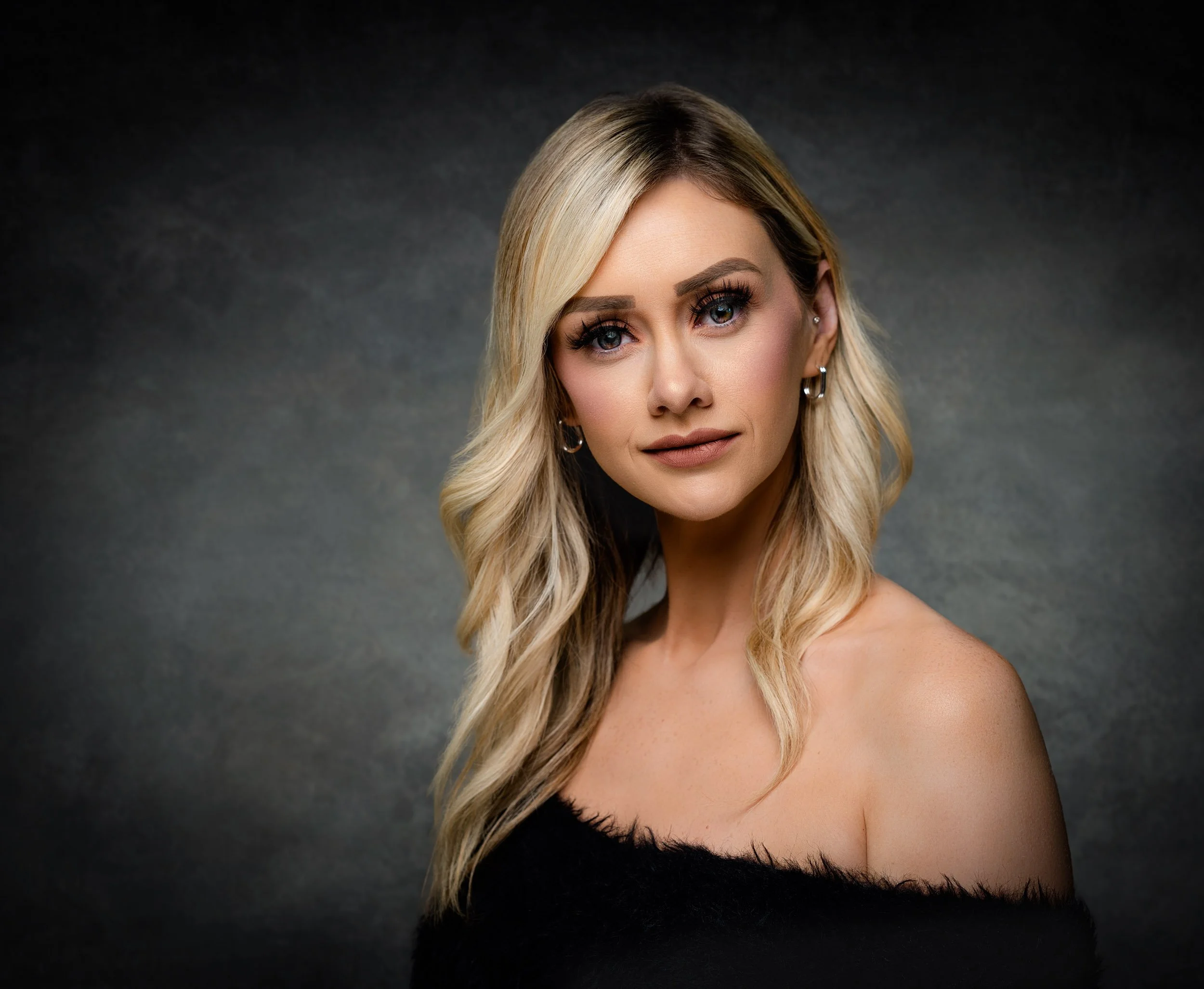 Portrait of a woman with long blonde wavy hair, wearing makeup, earrings, and a black off-shoulder top, against a dark textured background.