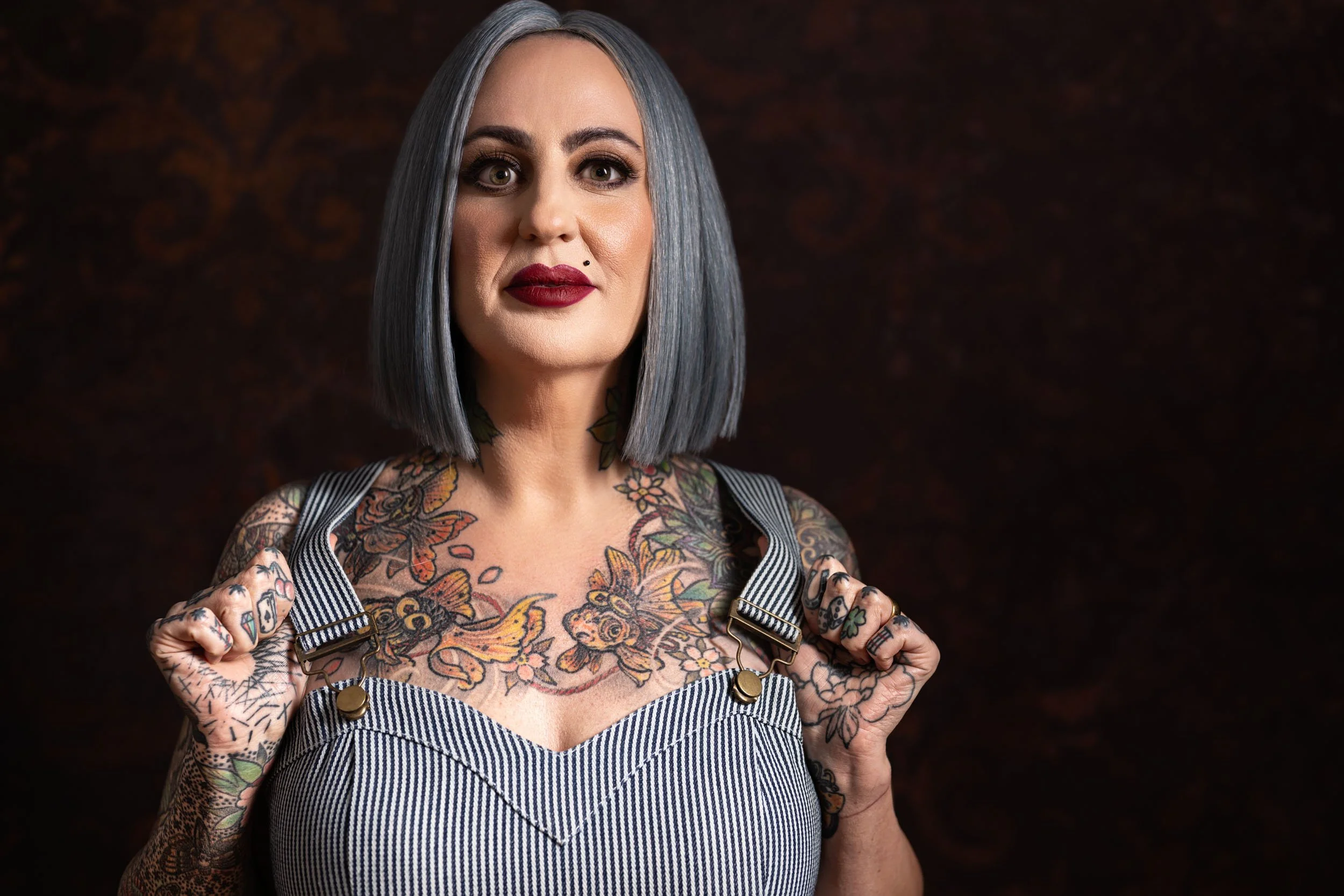 A woman with shoulder-length gray hair, dark eye makeup, and red lipstick, wearing a striped top and tattoos on her arms and chest, holding the straps of her top with both hands, against a dark background.