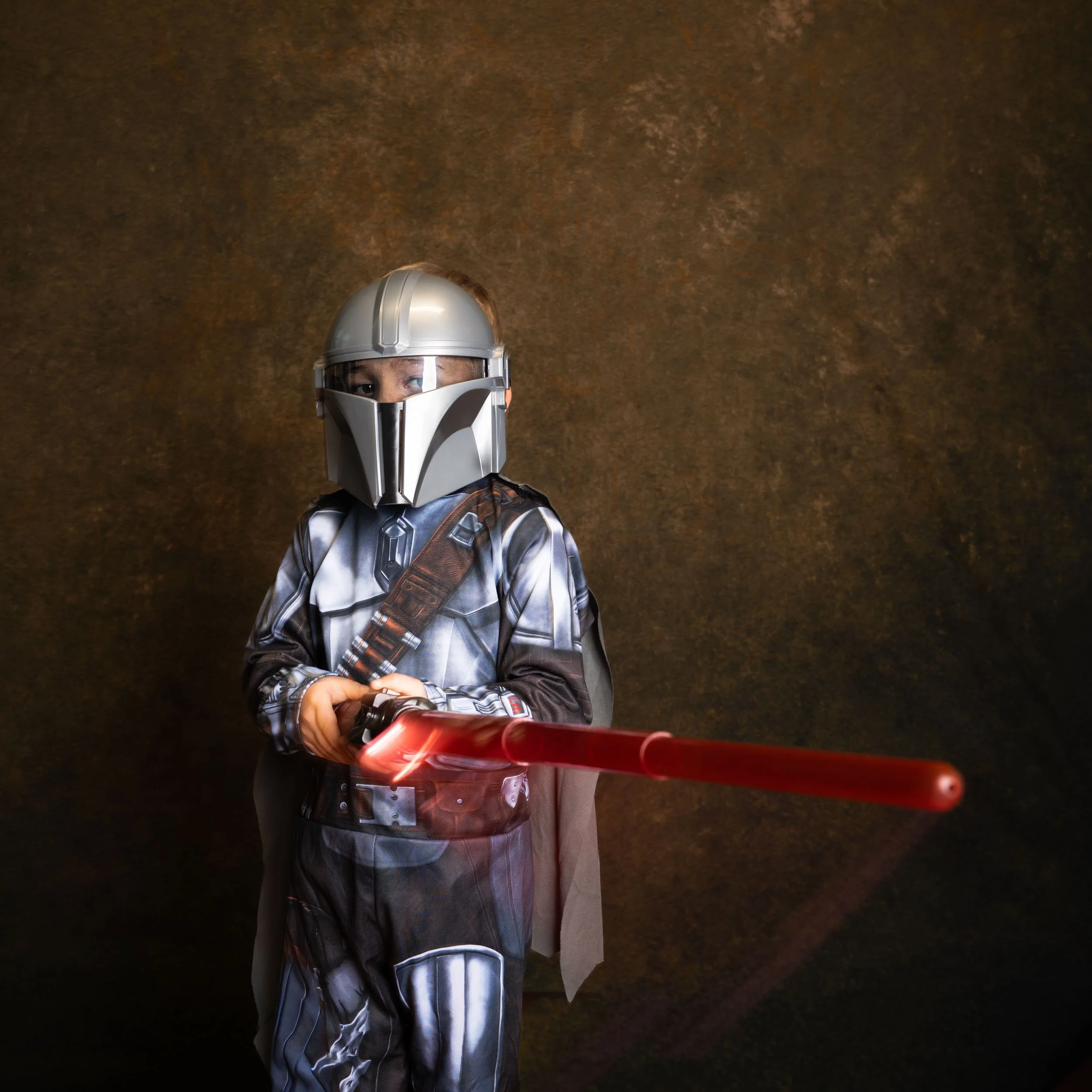 Child dressed as a Mandalorian from Star Wars, wearing a helmet and armor, wielding a red lightsaber.