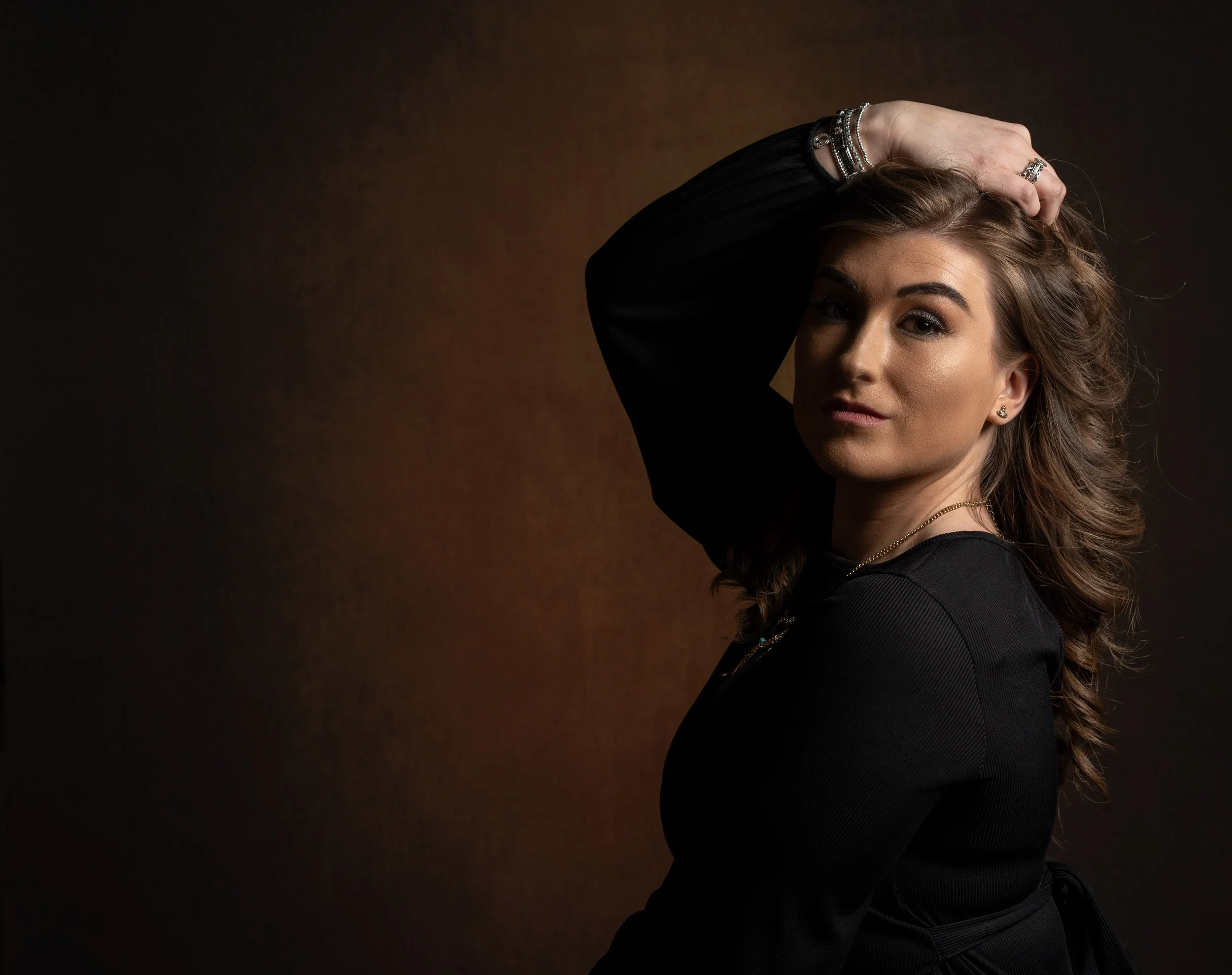 A woman with light skin and long wavy brown hair posing with one hand on her head against a dark gradient background.