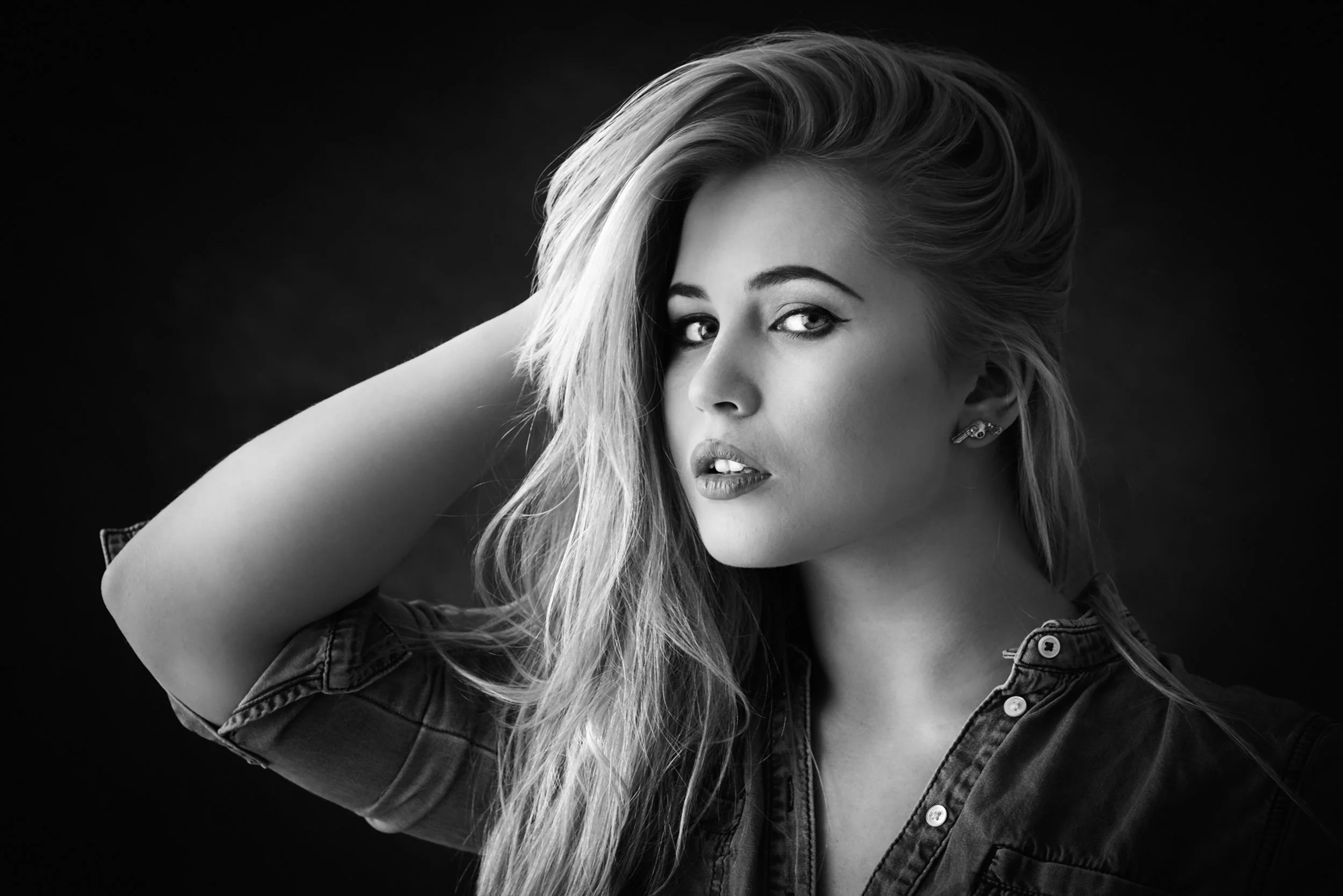 A black-and-white portrait of a woman with long, wavy hair, looking at the camera with her hand behind her head, wearing a denim shirt and earrings.