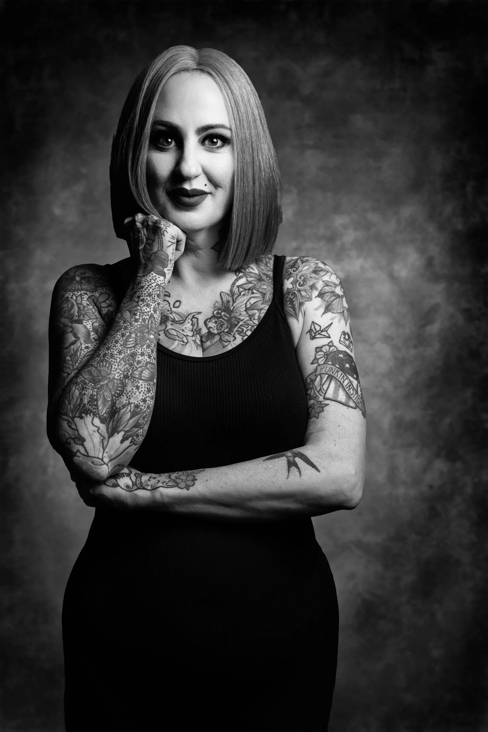 A woman with shoulder-length straight hair, tattoos on her arms and chest, wearing a black sleeveless top, posed with her left arm crossed and her right hand touching her face, against a textured dark background.
