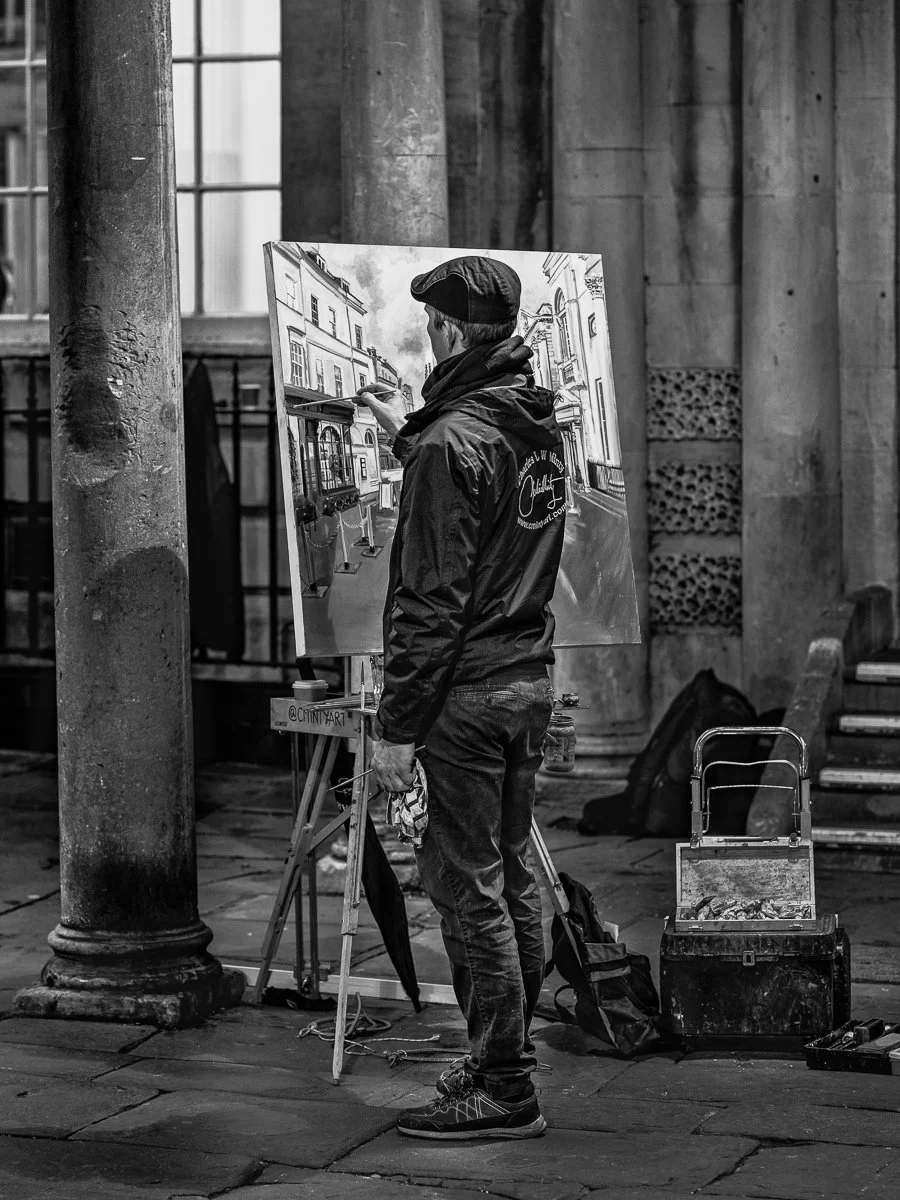 A man painting a cityscape on a canvas outside at night, standing next to a street lamp and his painting supplies.