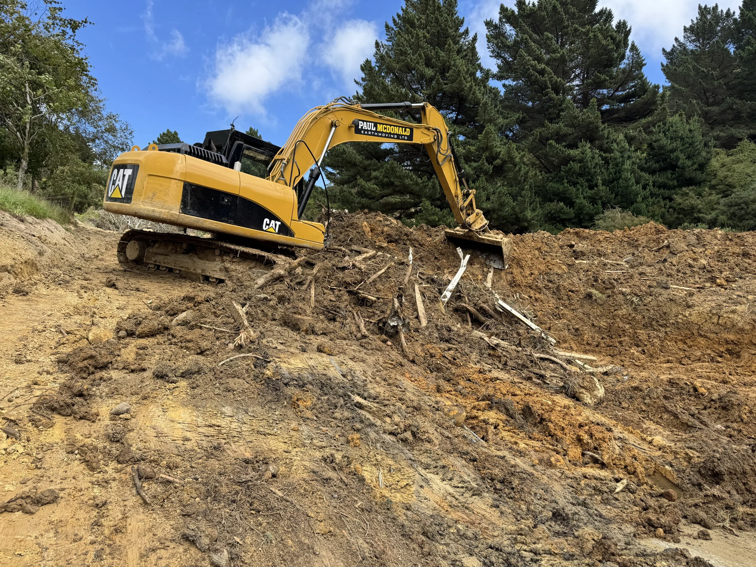 A yellow Caterpillar excavator working on a dirt hillside with trees and a partly cloudy sky in the background.