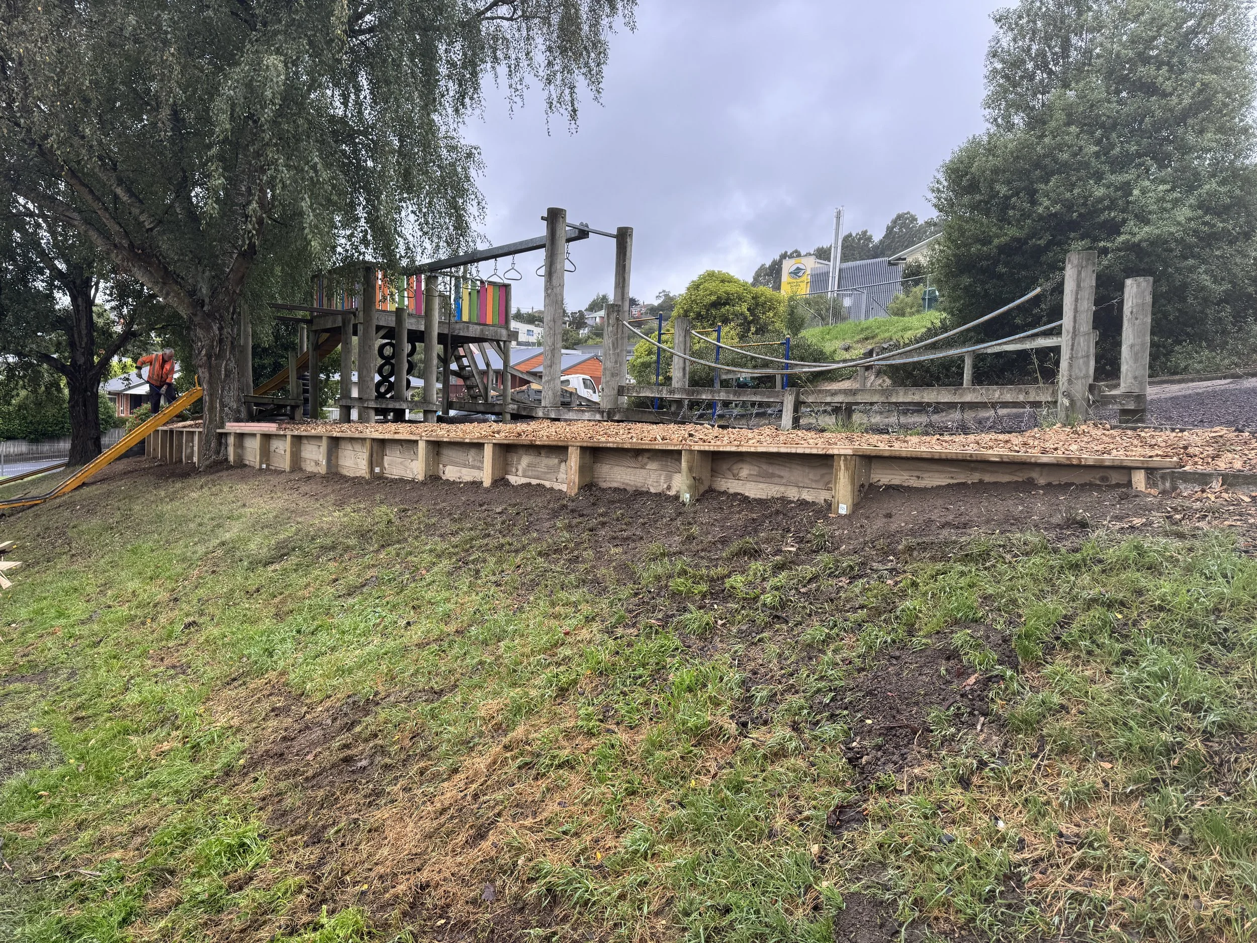 Construction of a new wooden ramp at a playground, with trees and construction worker in the background under an overcast sky.