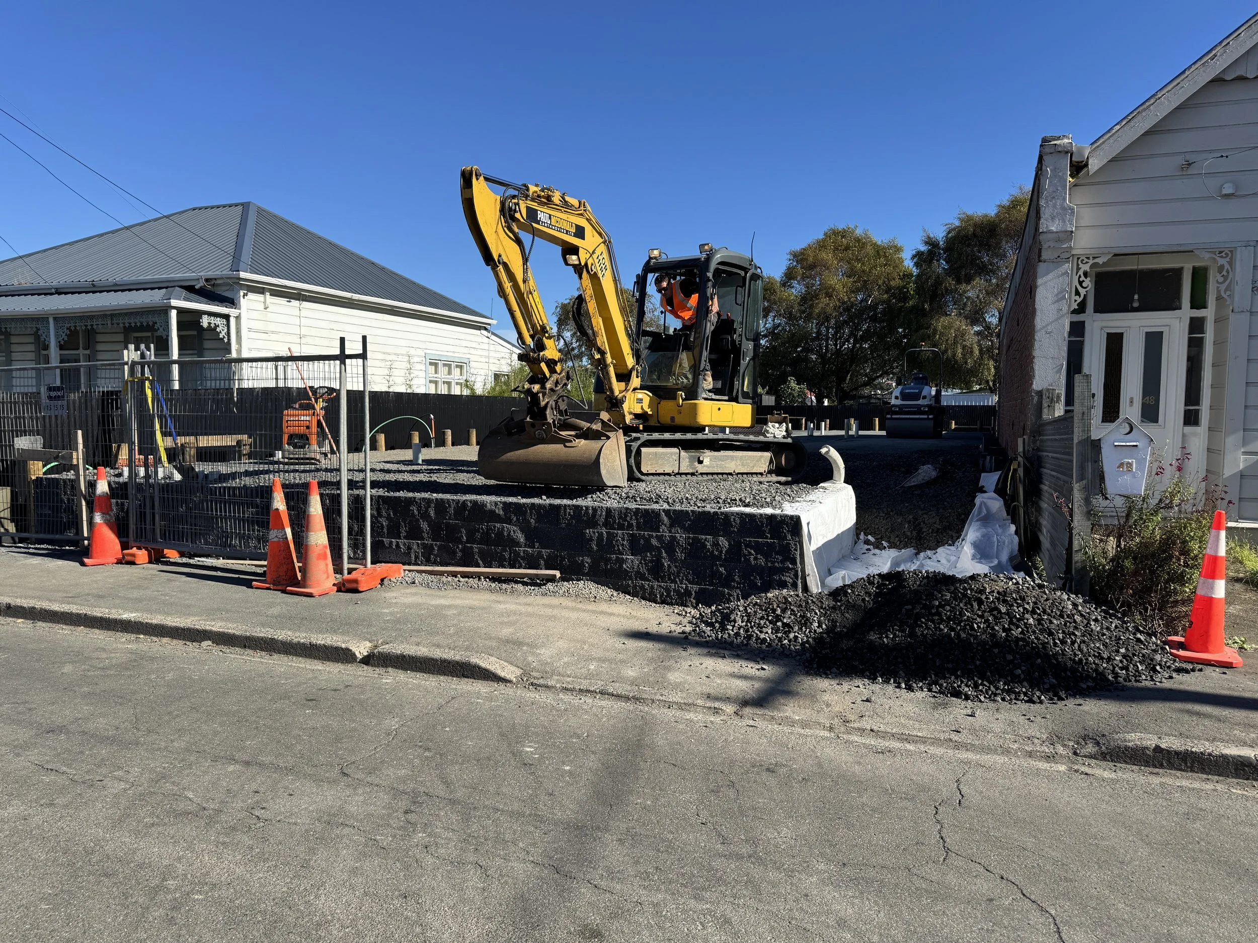 Construction site with a yellow excavator on a raised foundation, surrounded by orange safety cones and fencing, next to a residential house on a paved street.