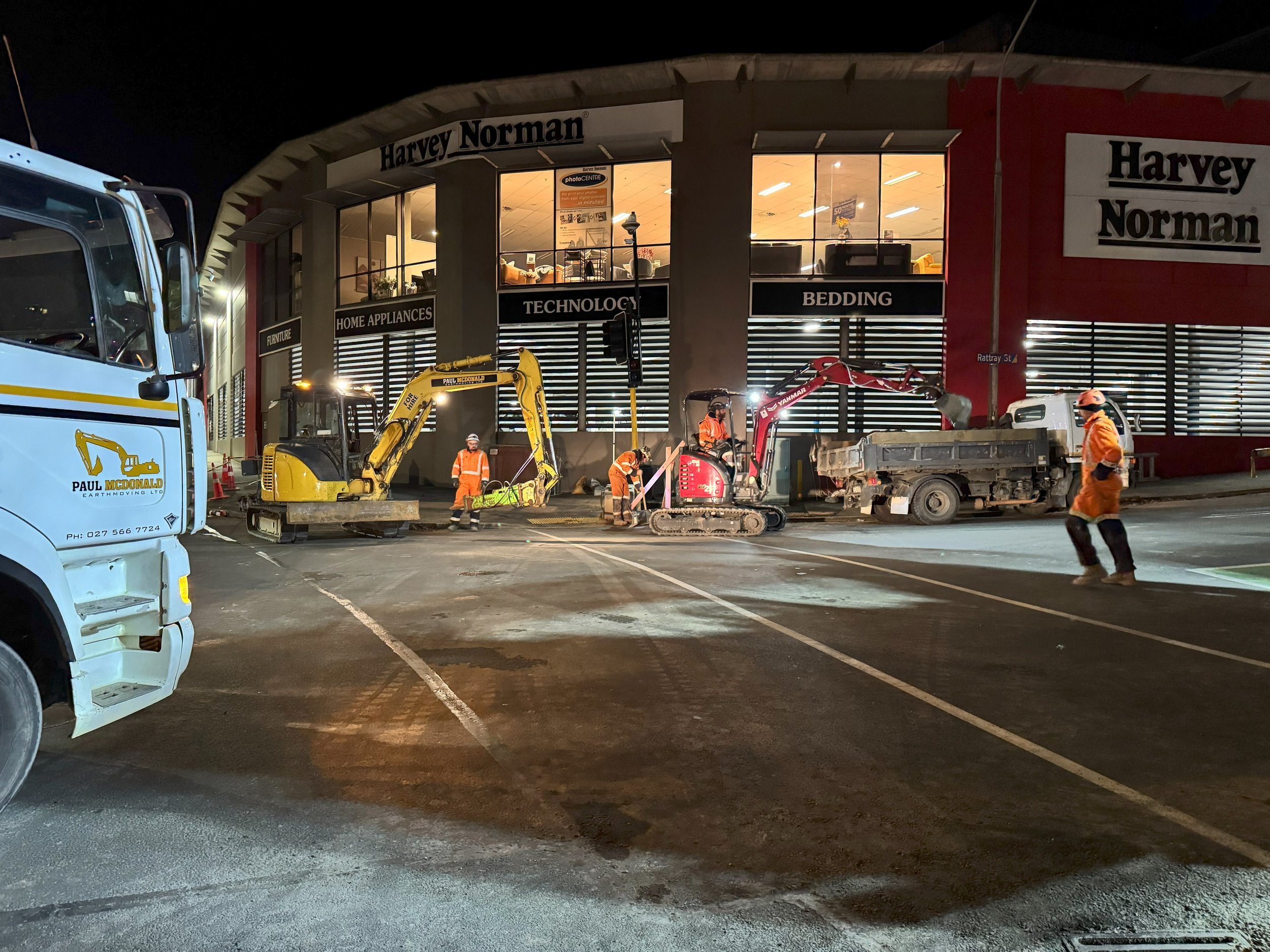 Nighttime construction scene outside Harvey Norman store with workers operating mini excavators and a small truck, several workers in orange safety gear, and a large white truck nearby.