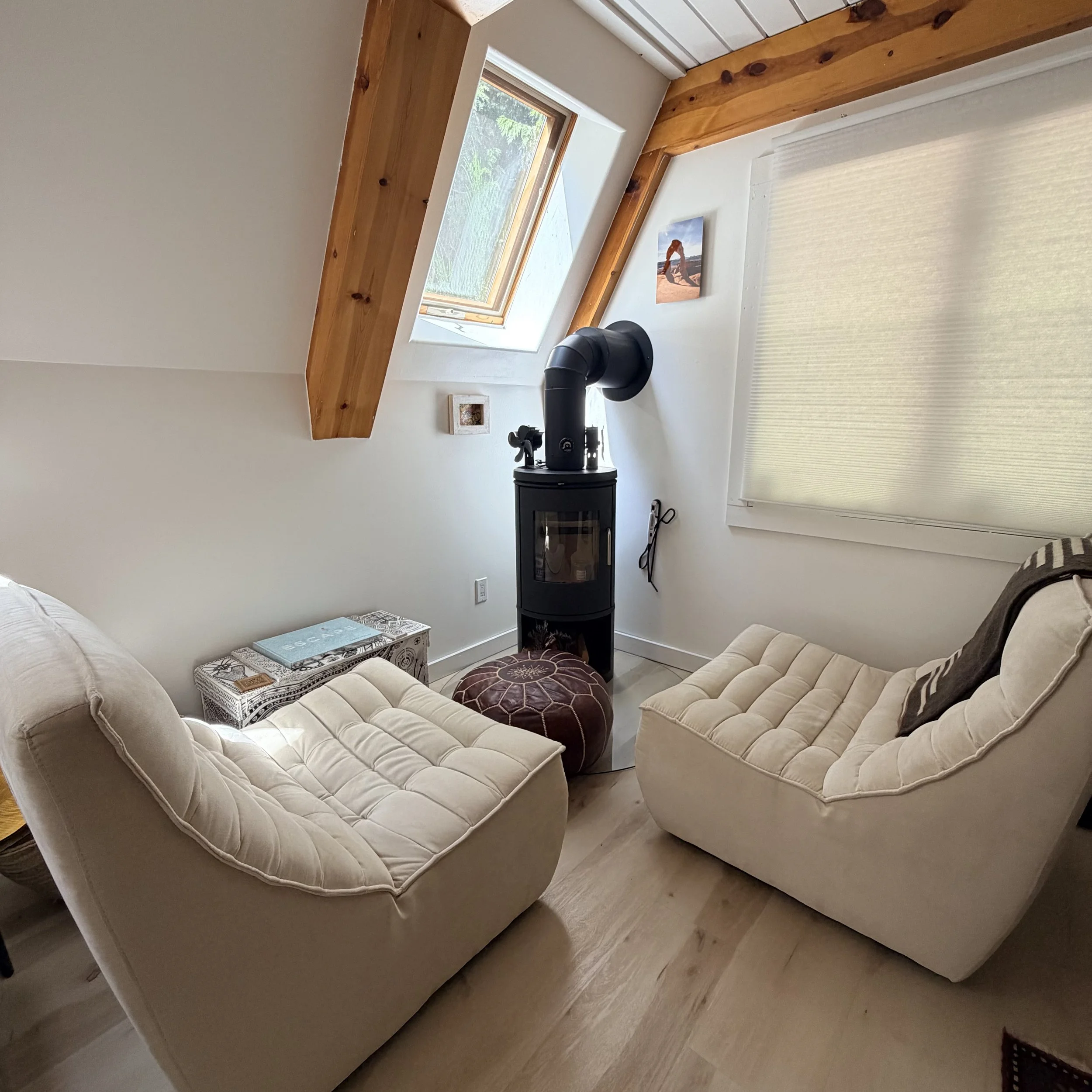 A cozy seating nook with beige sectional sofa, small side table with books, wood stove, window, and light-colored curtains where Stephanie Schuh, Life Coach in Carnation, WA and online, meets with her clients.