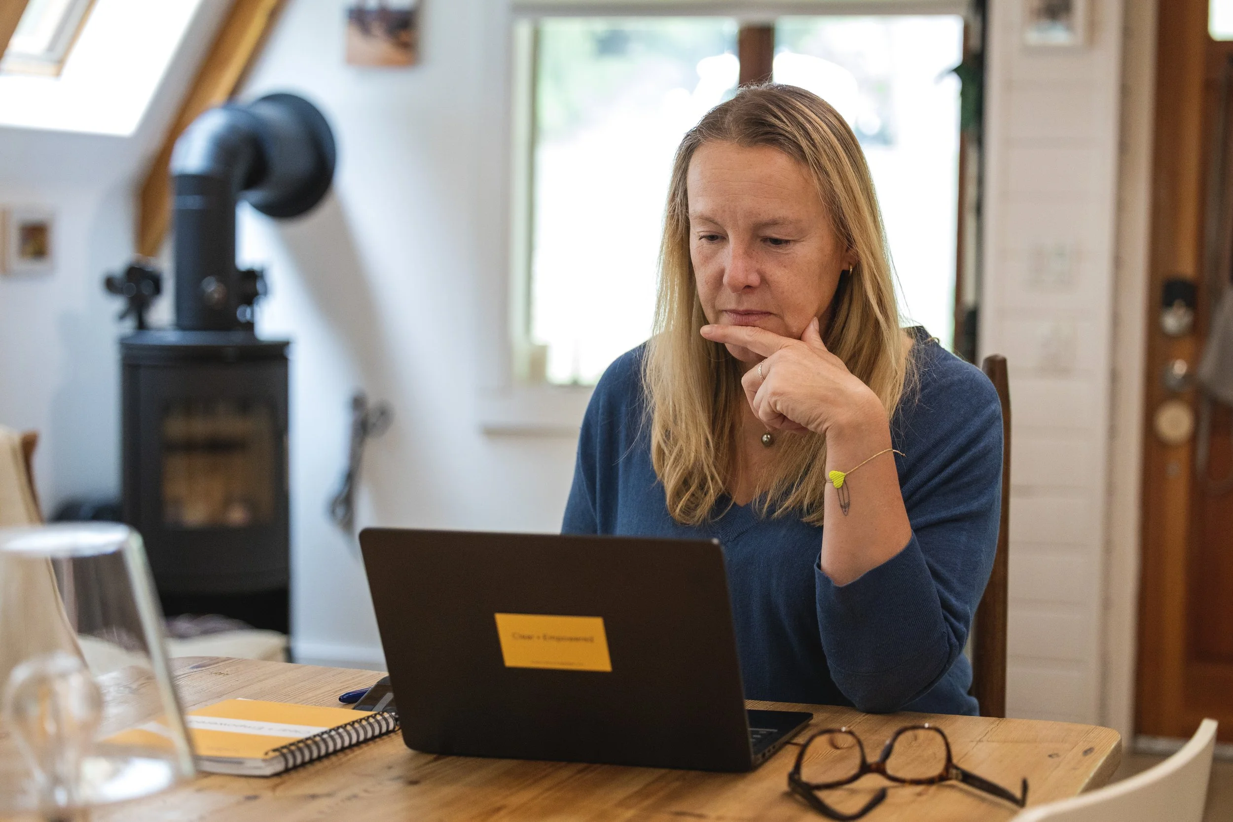 A photo of Stephanie Schuh, Life Coach in Carnation, WA and online, wearing a blue shirt, sitting at a wooden table, looking at a laptop, with her hand on her chin, in a cozy room with large windows, and a wood stove in the background.
