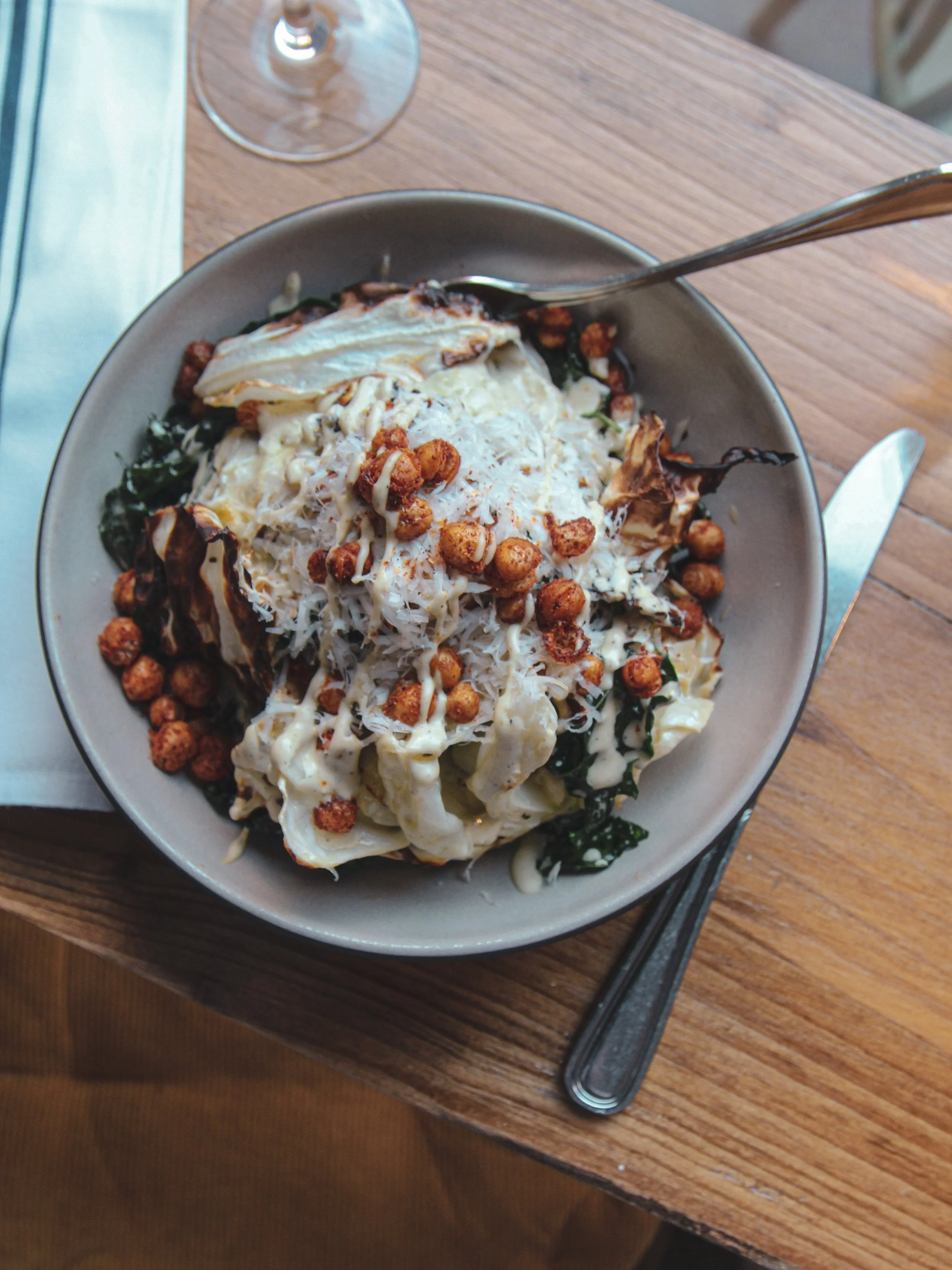 A vegan Caesar salad with lettuce, spinach, spicy roasted chickpeas, and grated cheese, served on a wooden table with a fork and knife.
