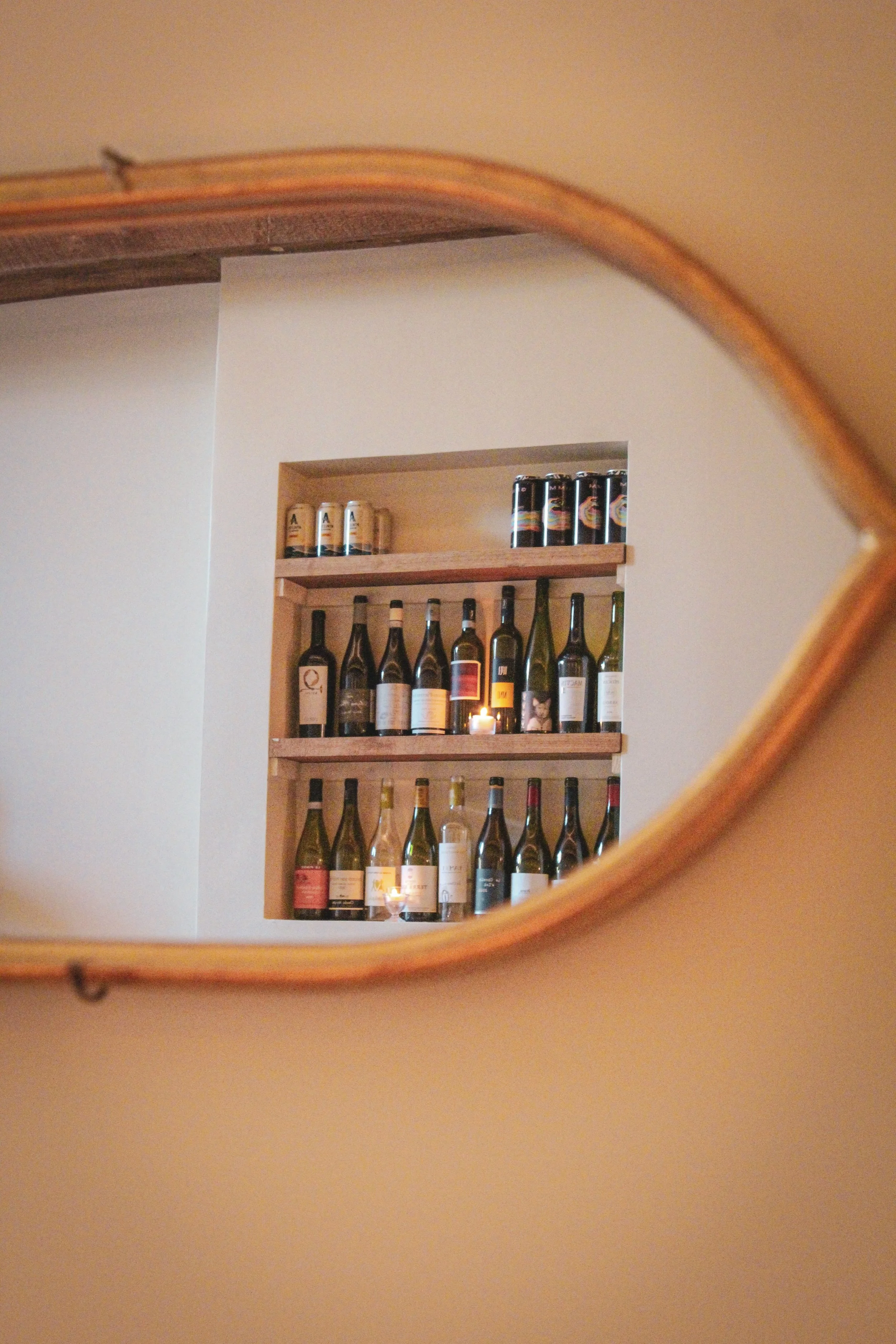 A collection of wine bottles and cans on wooden shelves, as seen through a mirror
