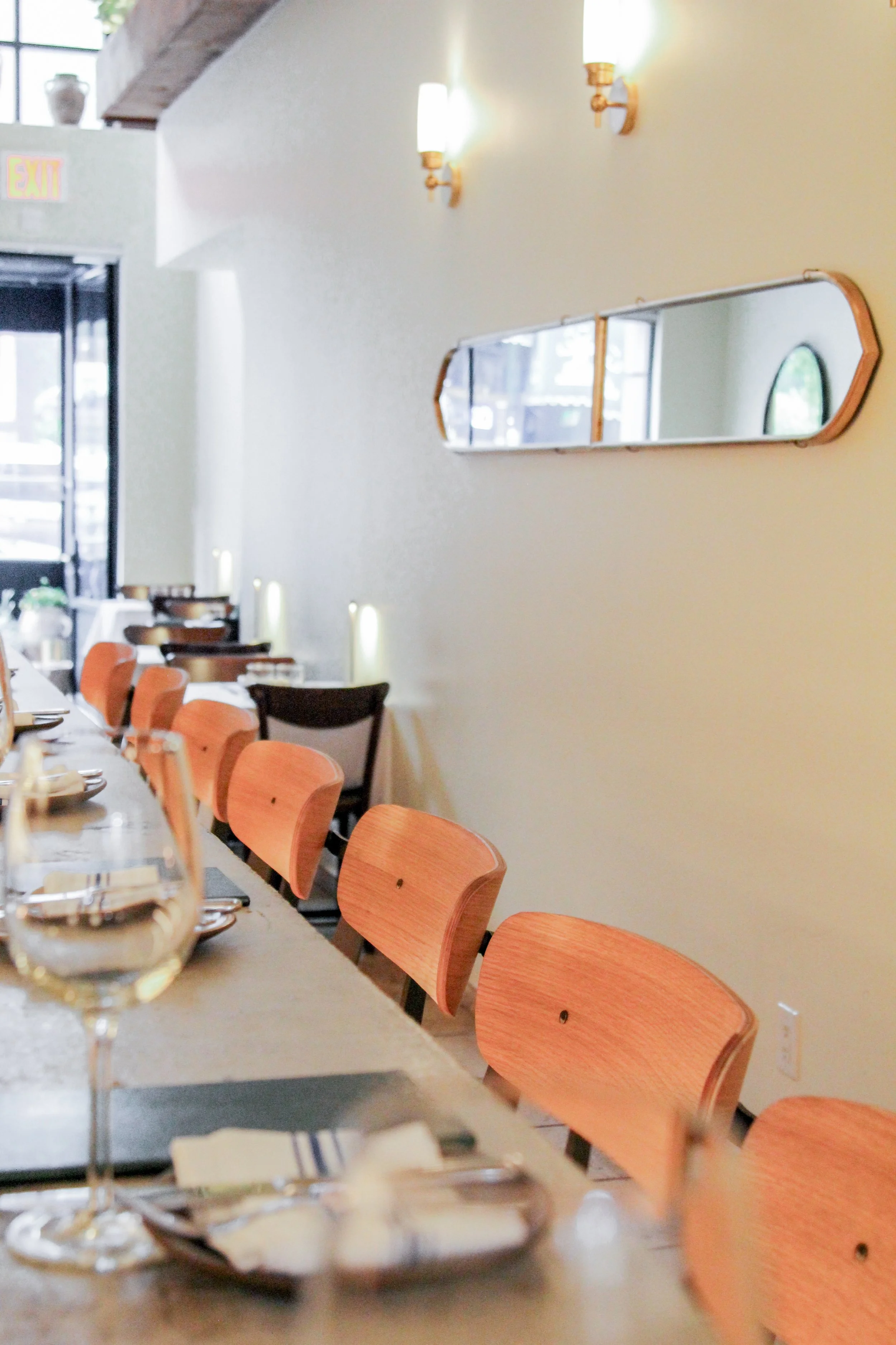 An indoor bar area with high wooden chairs, glassware, plates, and utensils. There are two wall-mounted lights on a beige wall, and a mirror is hung above the chairs. A door with glass panels is visible in the background, letting in natural light.