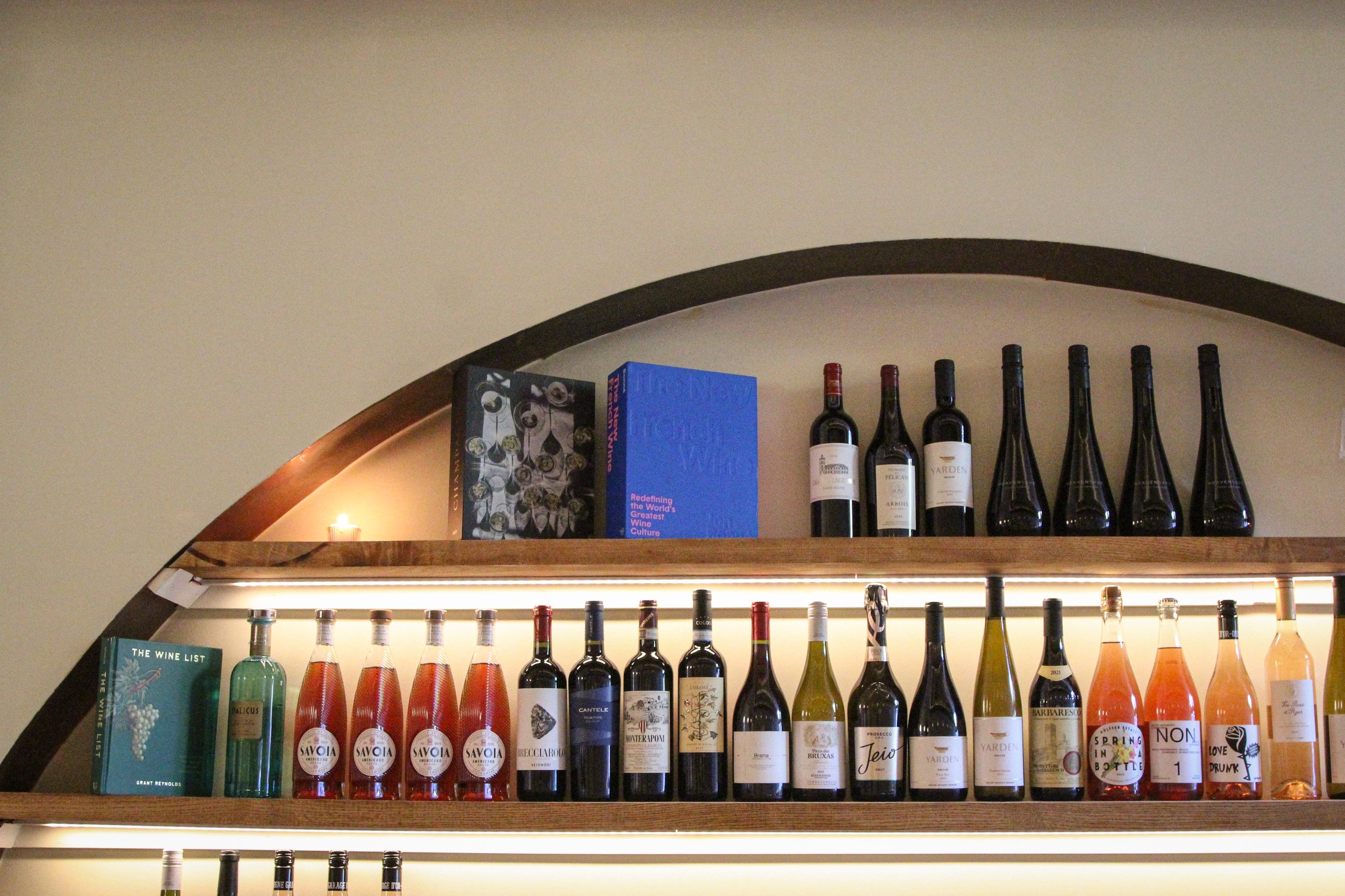 A wooden wine shelf displaying assorted wine bottles and books, with a candle in the background.