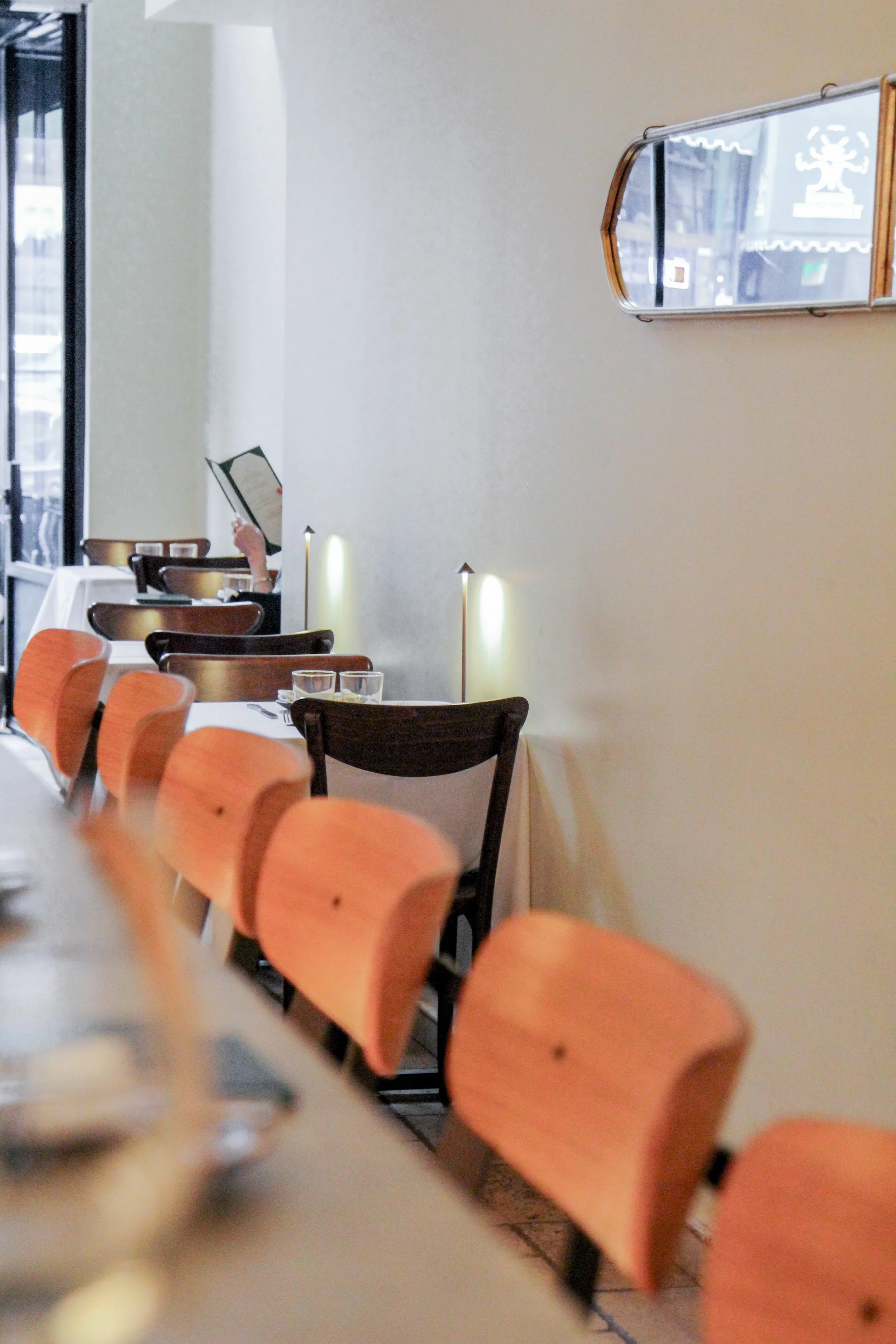 Bar area with wooden chairs, a person reading a menu in the background, and a wall-mounted mirror reflecting the street outside.