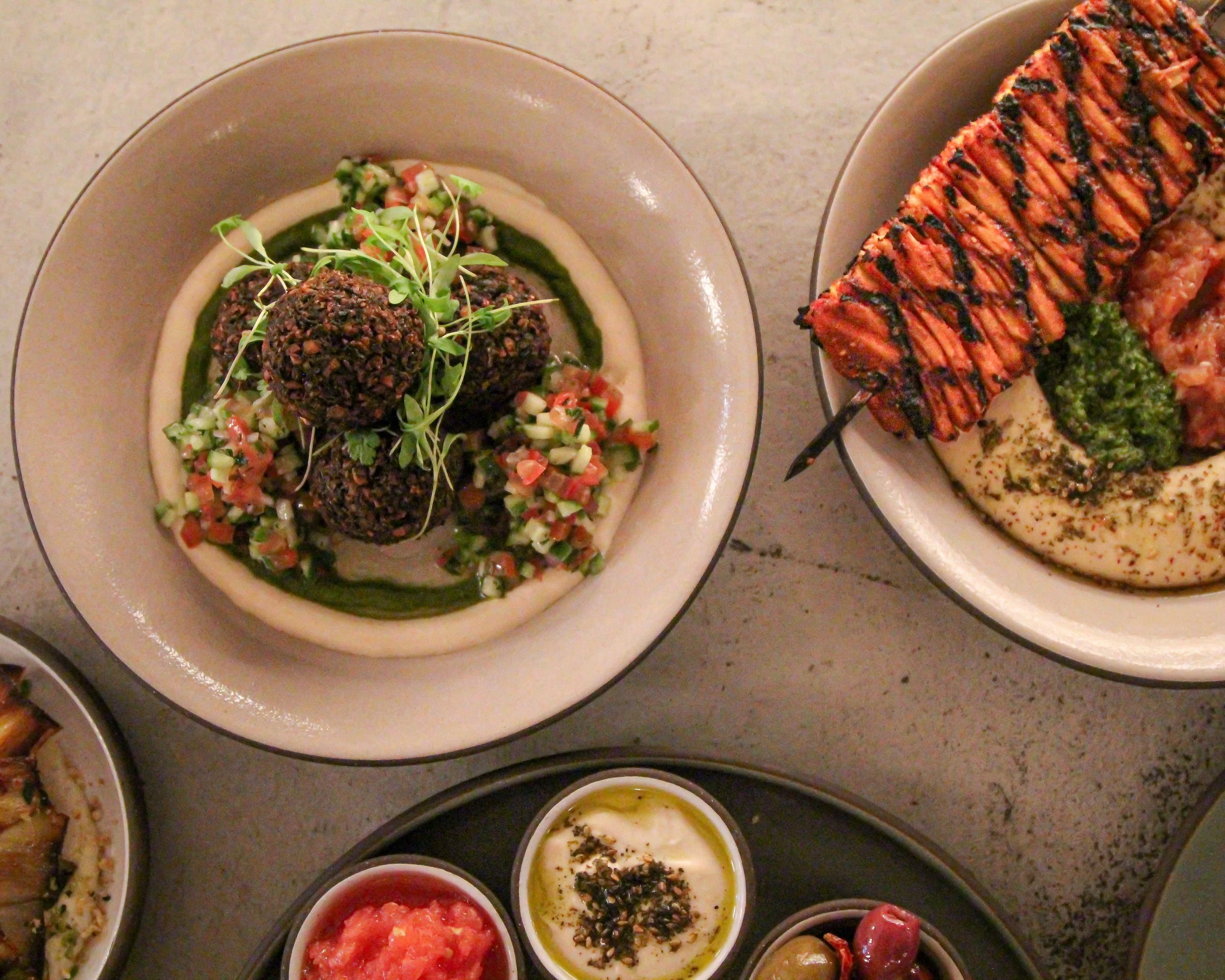 Part of a table with assorted dishes including a plate with falafel balls garnished with sprouts, chopped vegetables, and green sauce, and a plate with shawarma topped with herbs and drizzled with balsamic glaze.