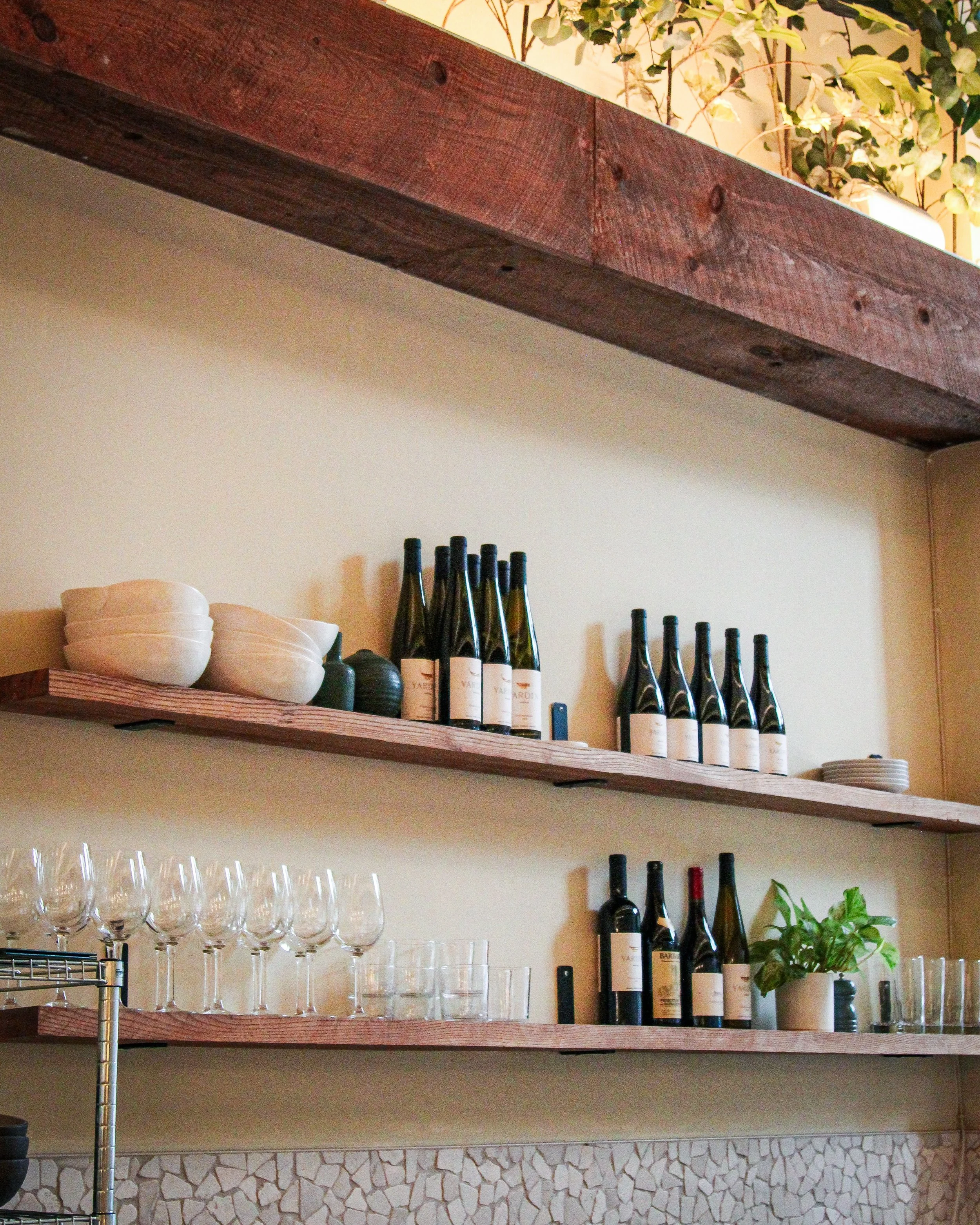 Wooden shelves with white plates, wine bottles, and empty wine glasses in a restaurant or bar setting, with some greenery and a textured stone wall below.
