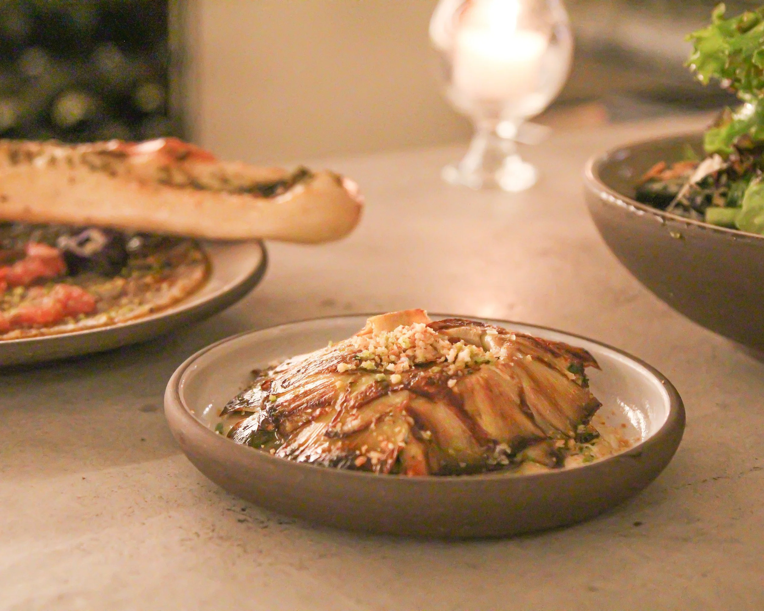 Close-up of a bowl of deconstructed artichoke garnished with chopped nuts on a beige countertop, with other dishes and a wine glass in the background.