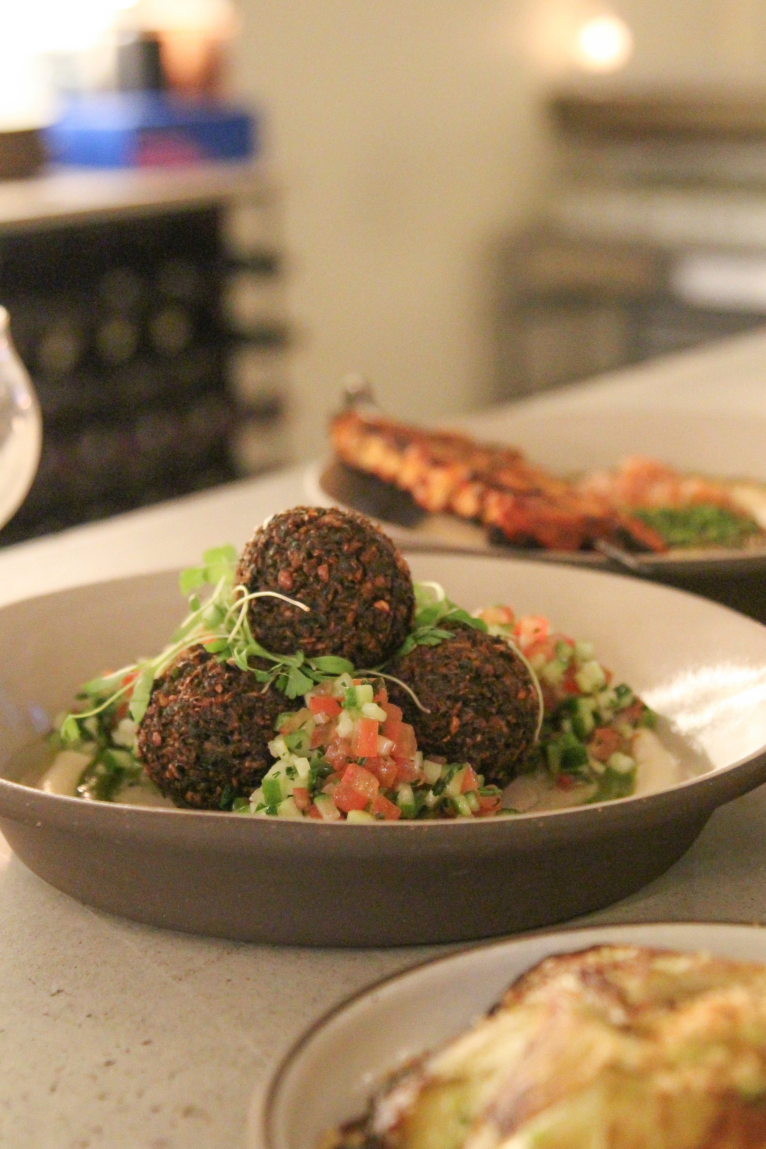 Close-up of a bowl of falafel balls topped with microgreens and served over tabbouleh salad with diced vegetables, on a kitchen counter. In the blurred background, there is shawarma on a black plate.