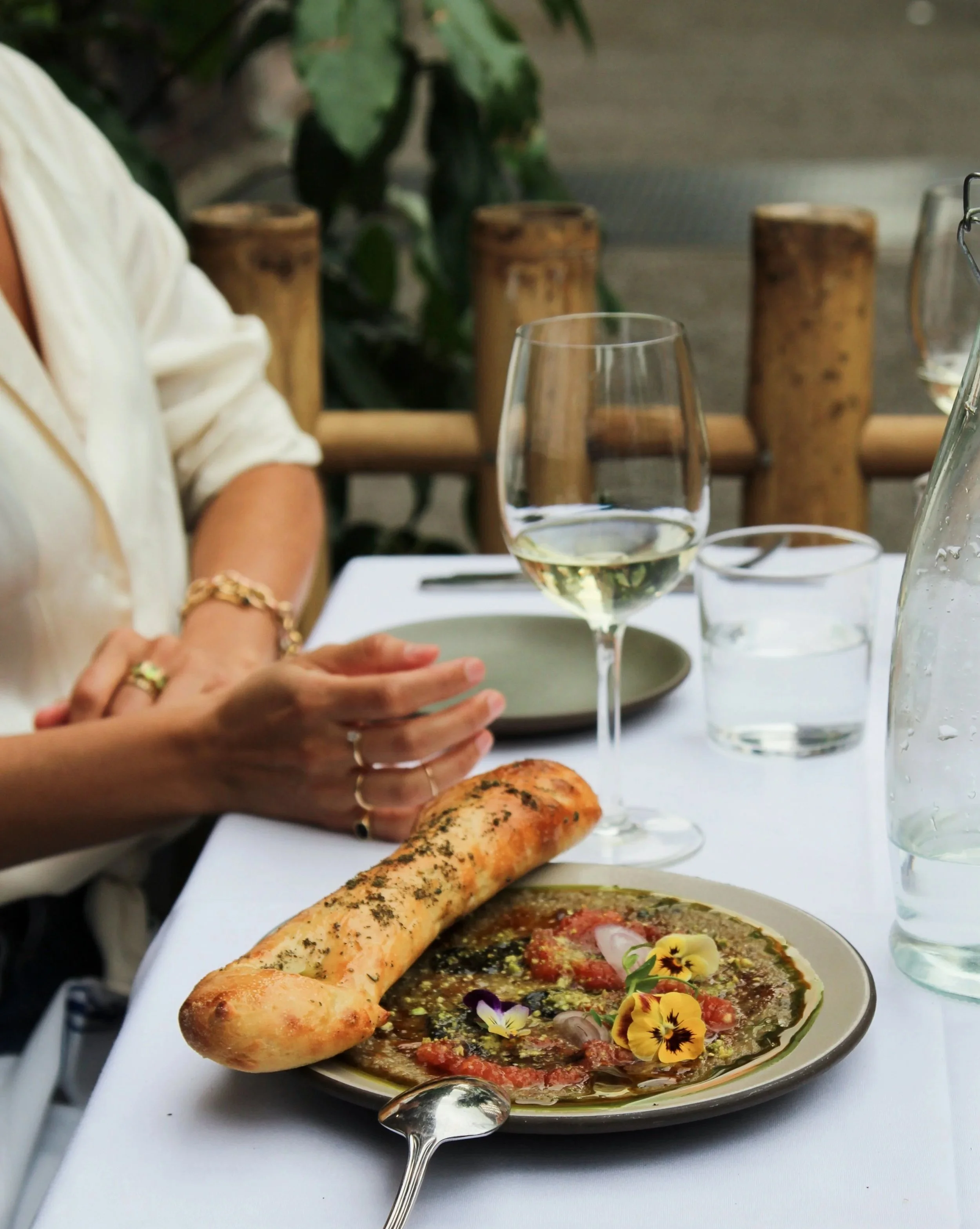 A restaurant table with a plate of eggplant garnished with edible flowers, a side of naan bread, a glass of white wine, and a glass of water. In the background, a person with jewelry sits across the table, with wooden decor and green plants visible.