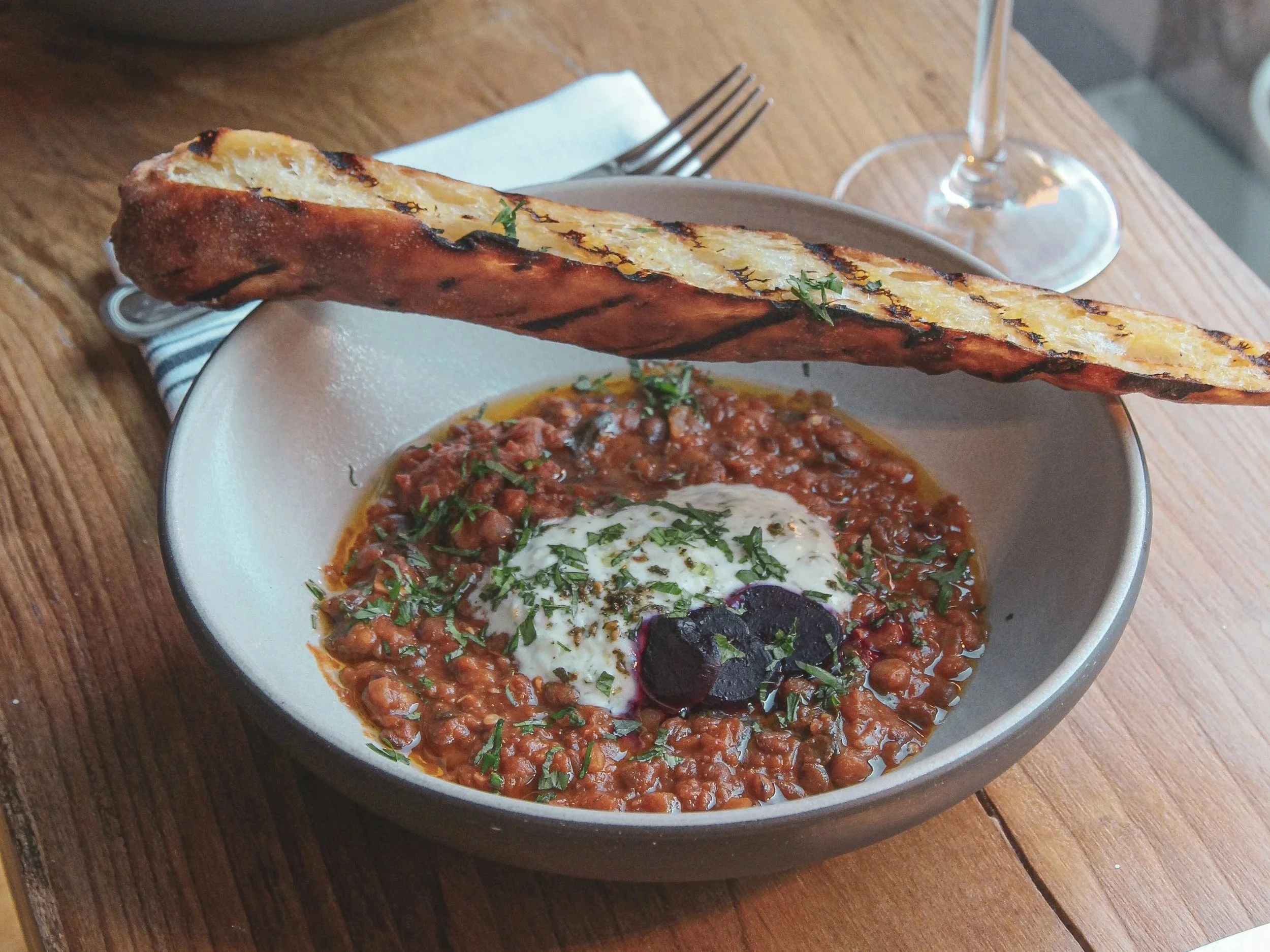 Bowl of chili garnished with chopped herbs and topped with a creme fraiche, on a wooden table with a fork and wine glass in the background.