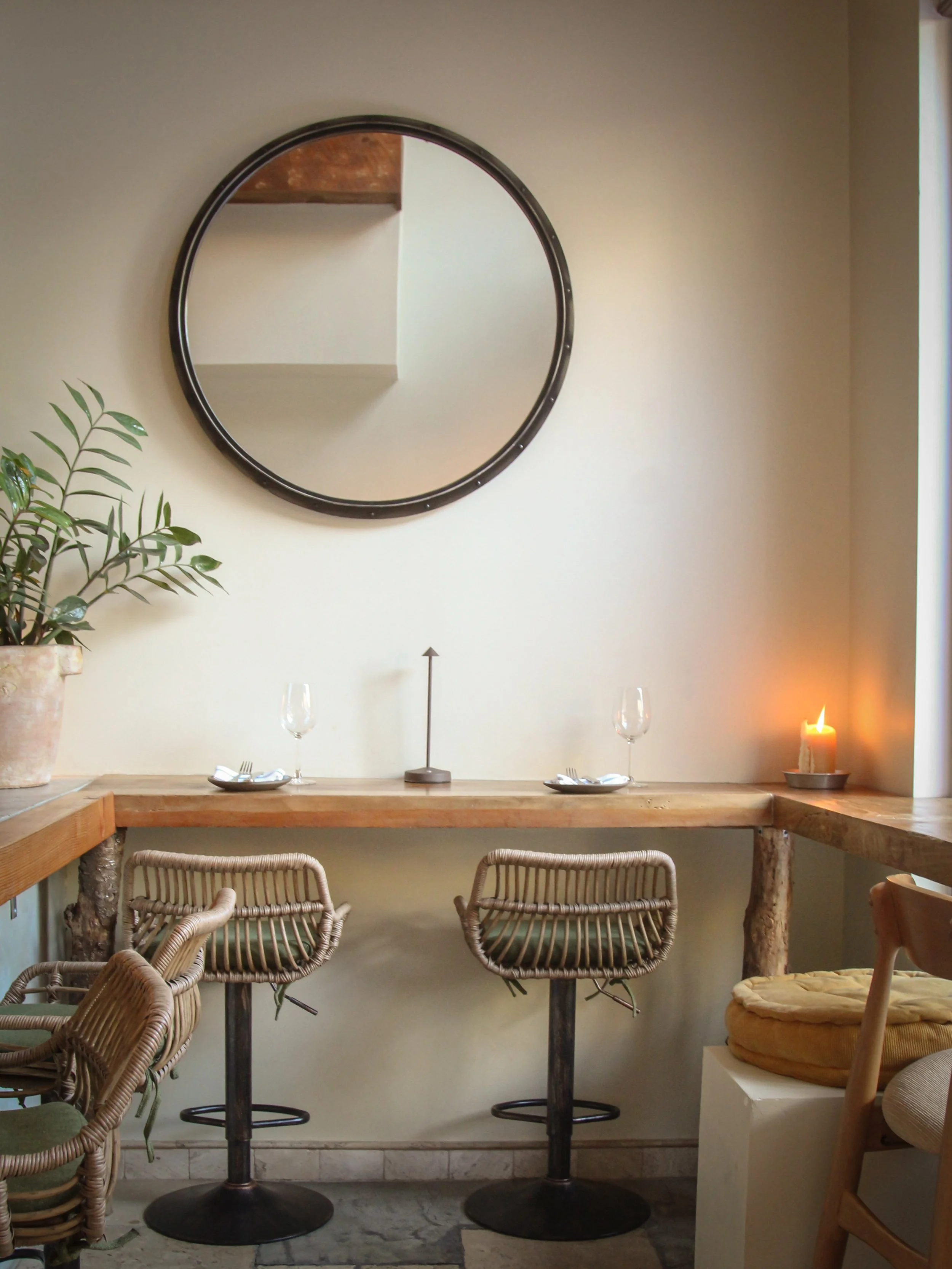A cozy dining nook with a wooden table and rattan chairs, decorated with a potted plant, a white candle, and a large round mirror on the wall.