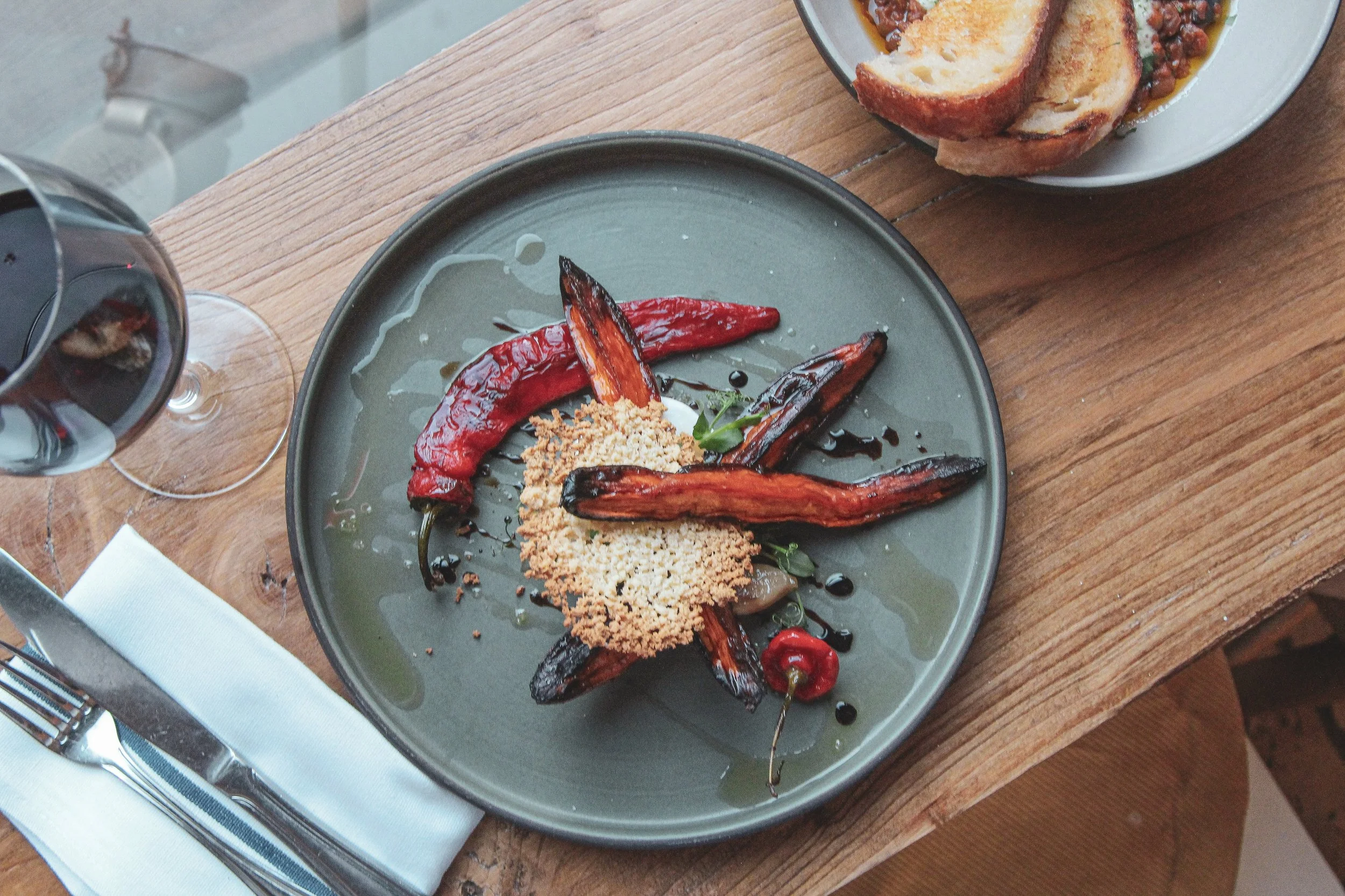 A plate of grilled red chili peppers with crisped vegan parmesan cheese and marinated oils, accompanied by a glass of red wine and bread with beans and sauce on a white dish, on a wooden table.