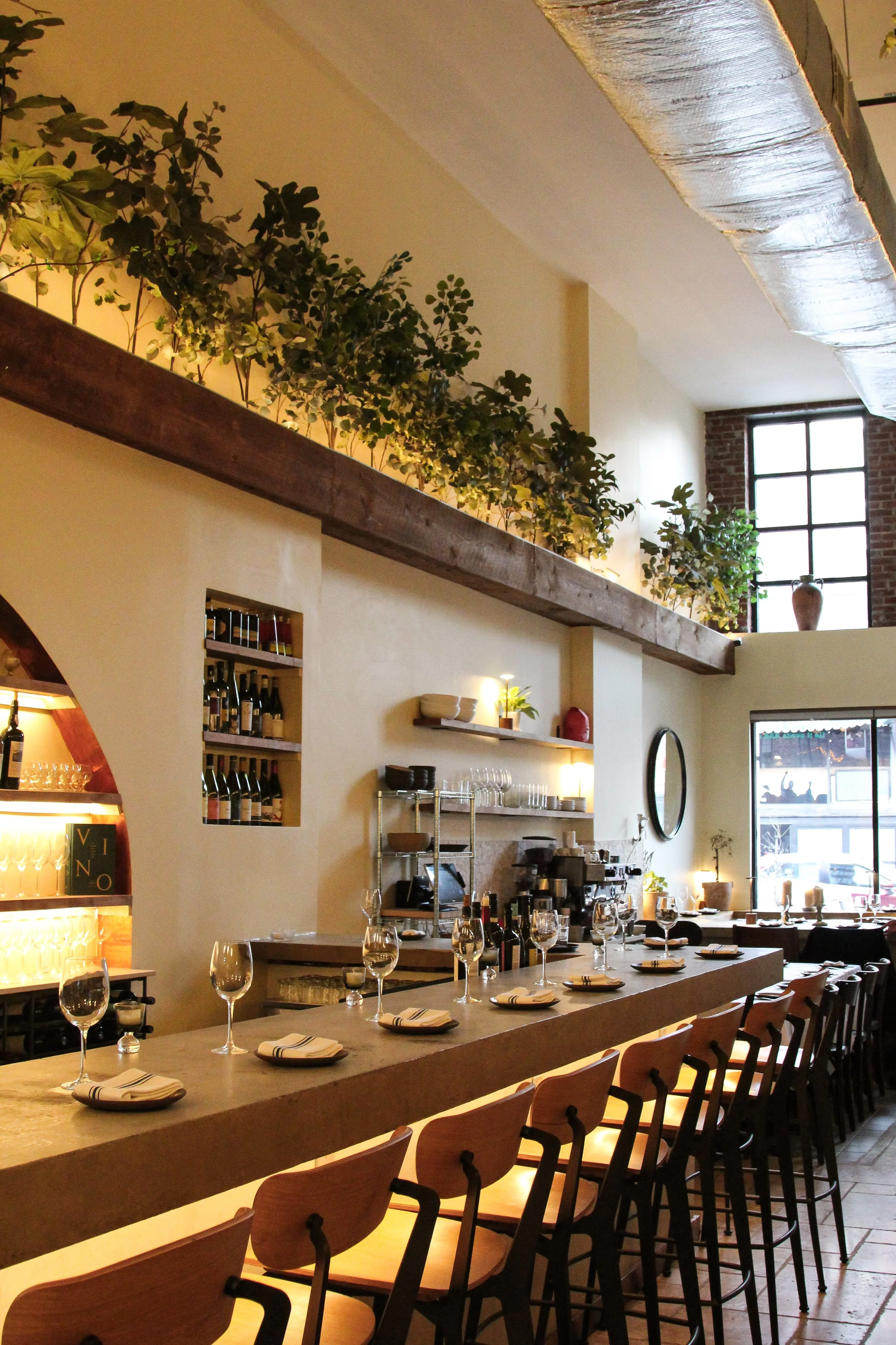 A cozy indoor restaurant with wooden chairs and tables set for dining, featuring wine glasses and napkins, with a kitchen area in the background and a large window bringing in natural light.