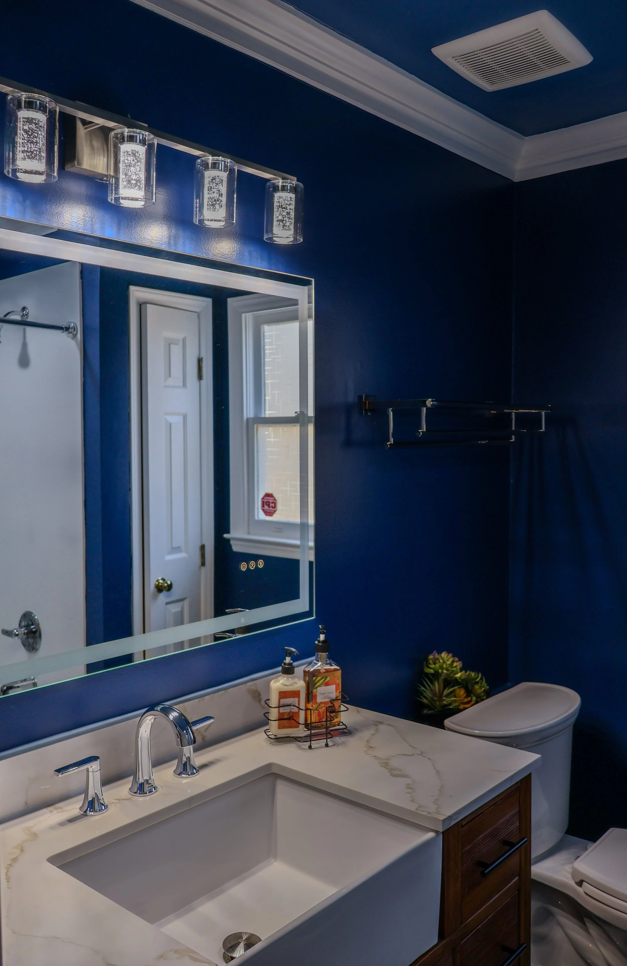 A bathroom with dark blue walls, a white marble countertop with a sink, a mirror, and a light fixture with four glass shades. There is a window reflecting in the mirror, a toilet, a small plant, and some lotion bottles on the counter.
