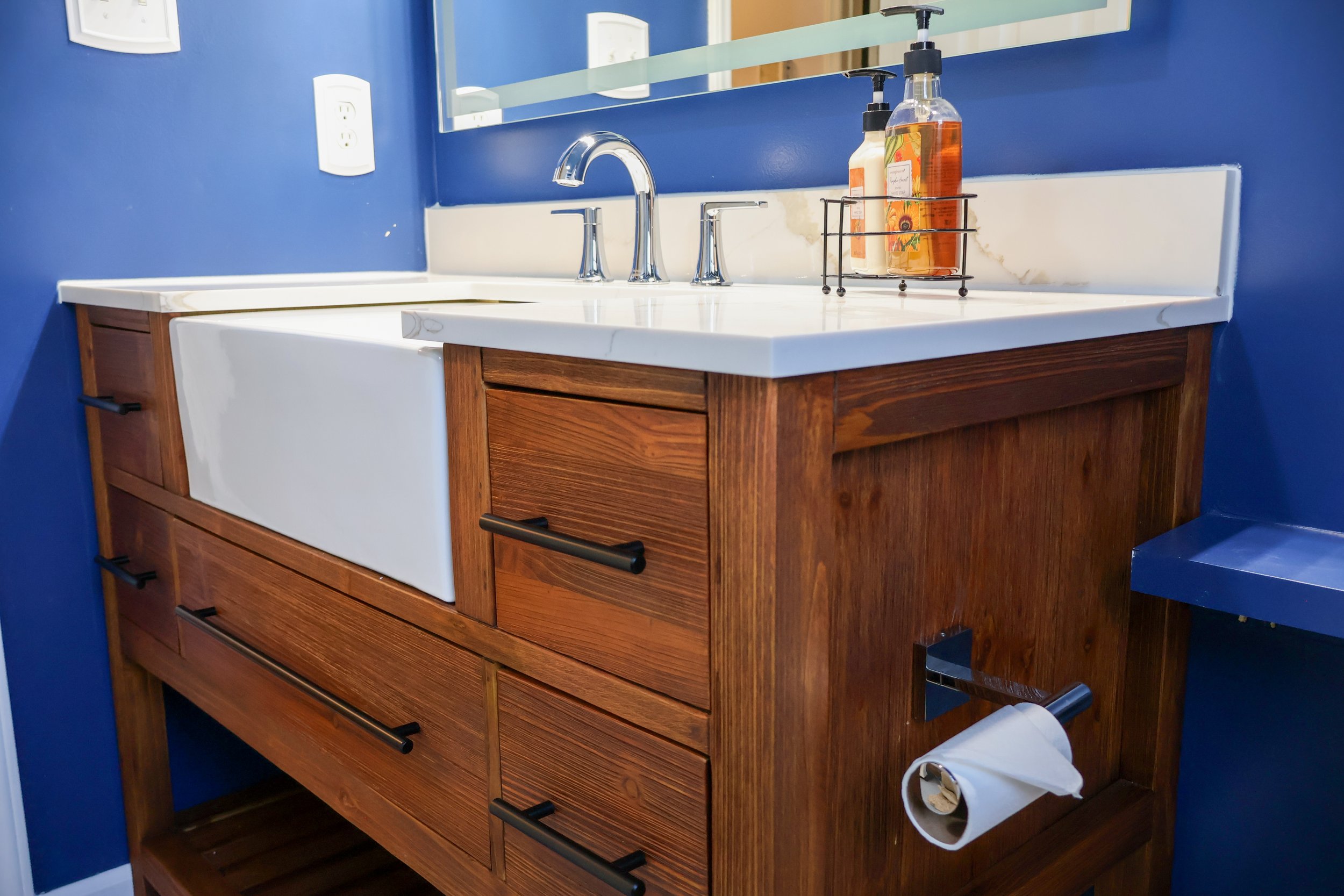 Wooden bathroom vanity with a white ceramic sink, chrome faucet, and a mirror above. There are soap and lotion bottles on a rack, a white wall socket, and a roll of toilet paper on a holder on the right side.
