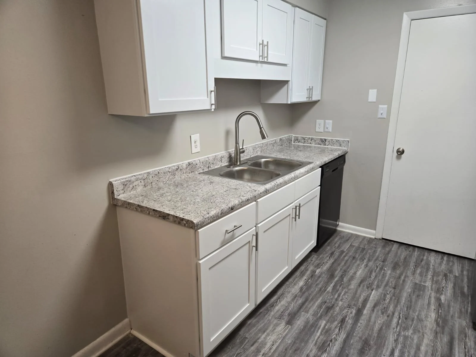 Small kitchenette with white cabinets, granite countertop, stainless steel double sink, black dishwasher, beige walls, and gray wood-look flooring.