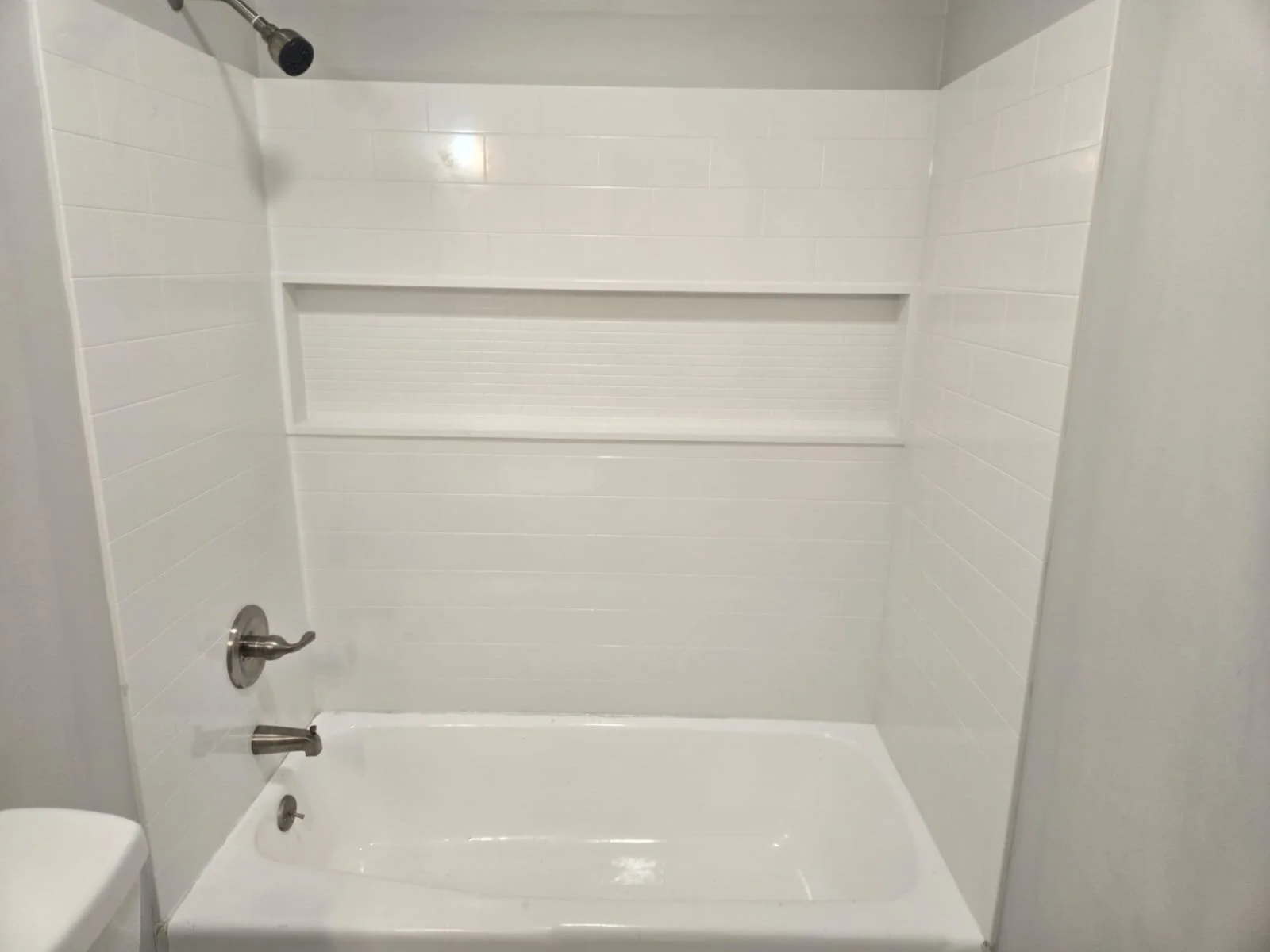 Empty white bathtub in a shower area with white tiled walls, a handheld showerhead, and a wall niche for toiletries.