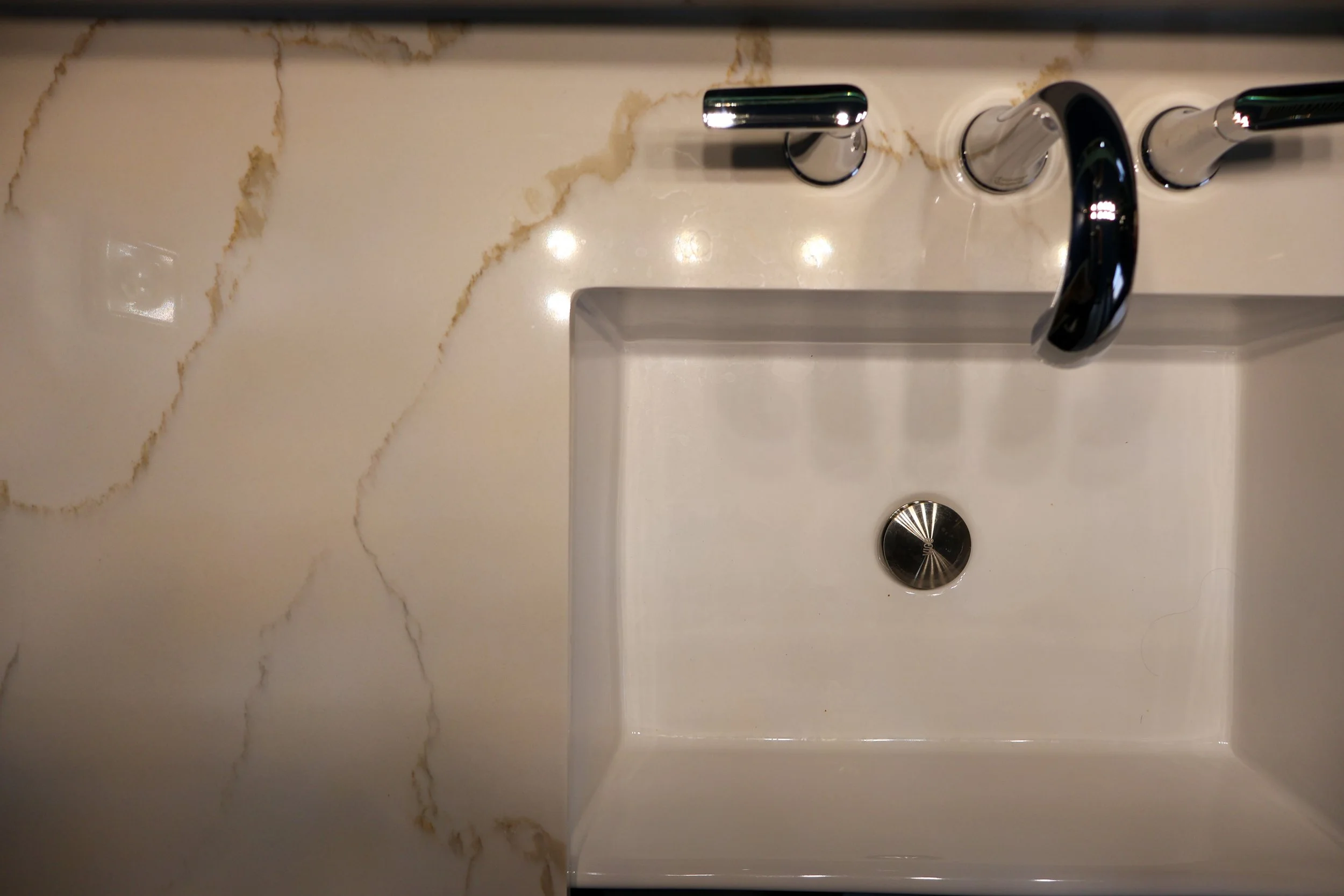 A white bathroom sink with a shiny chrome faucet and three handles, installed on a cream-colored marble countertop.