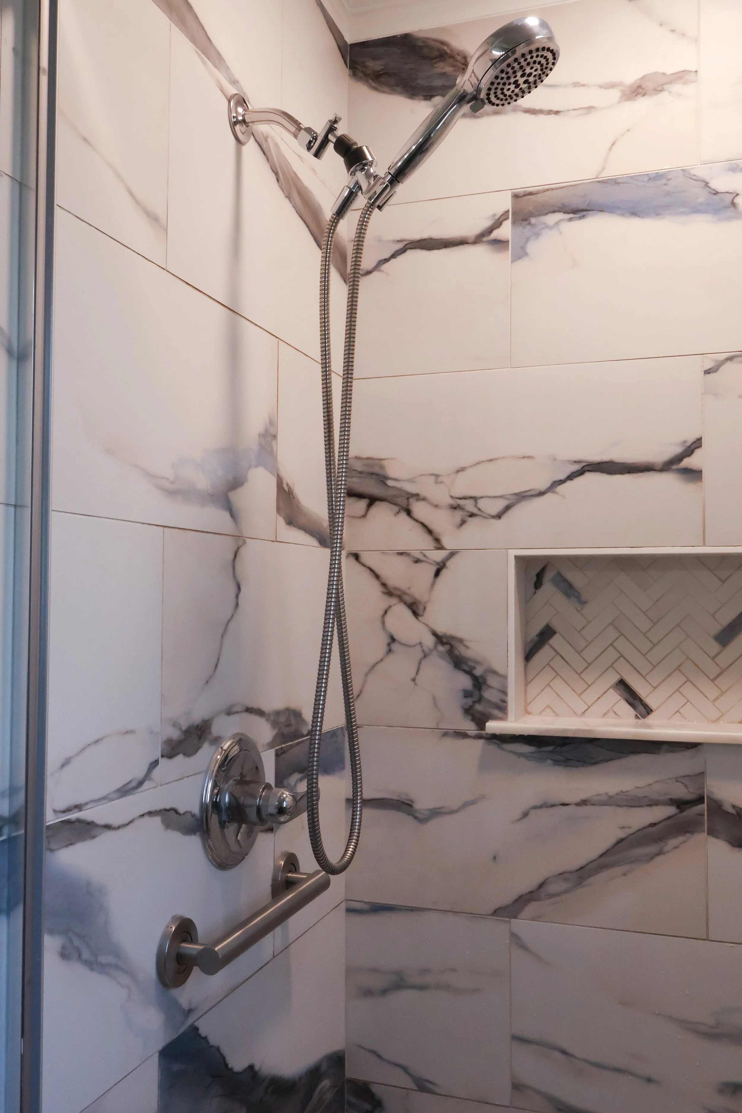 A walk-in shower with large marble-style tiles, a chrome showerhead, a handheld shower wand, and a built-in recessed shelf with a herringbone tile interior.