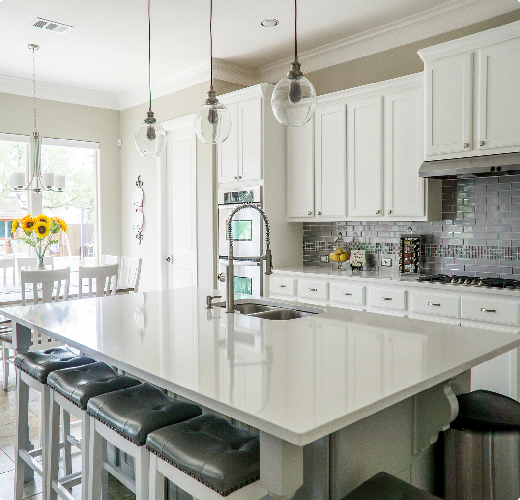 Modern kitchen with white cabinets, a large white island with black cushioned stools, pendant lights, gray tile backsplash, stainless steel appliances, and a dining area with a vase of sunflowers.