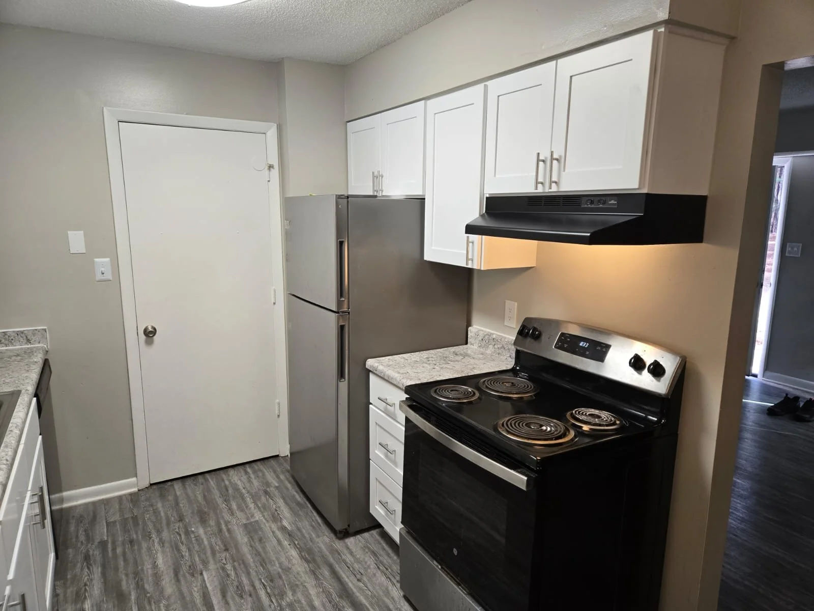 A kitchen with white cabinets, a stainless steel refrigerator, a black electric stove, a black range hood, light-colored countertops, and gray wood-look flooring.
