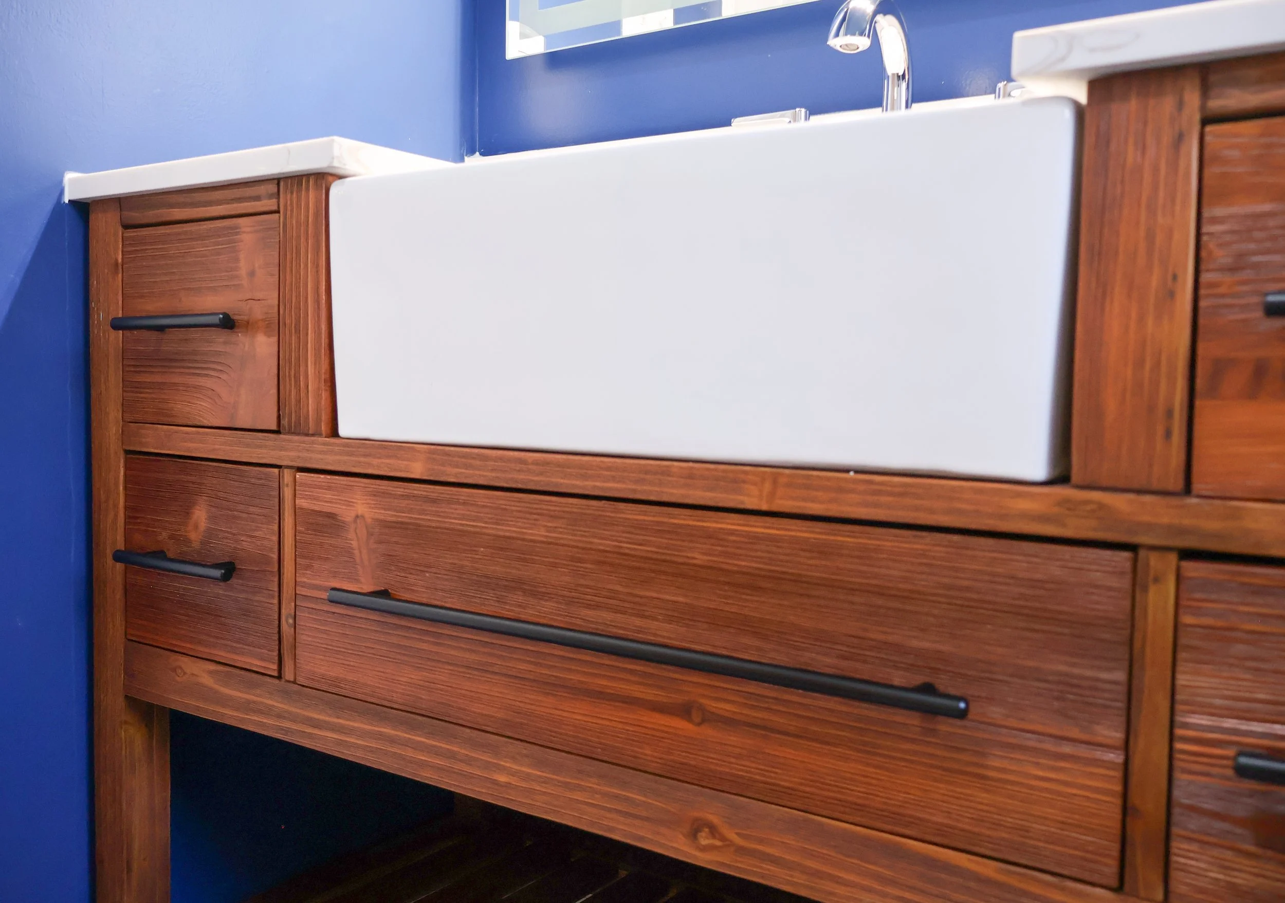 Bathroom sink with a white apron front, set in a wooden vanity cabinet with black handles, against a blue wall.