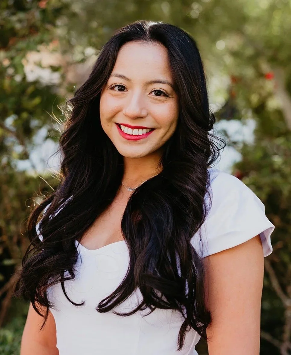A young woman with long, wavy black hair, smiling, wearing a white top, outdoors with blurred greenery background.