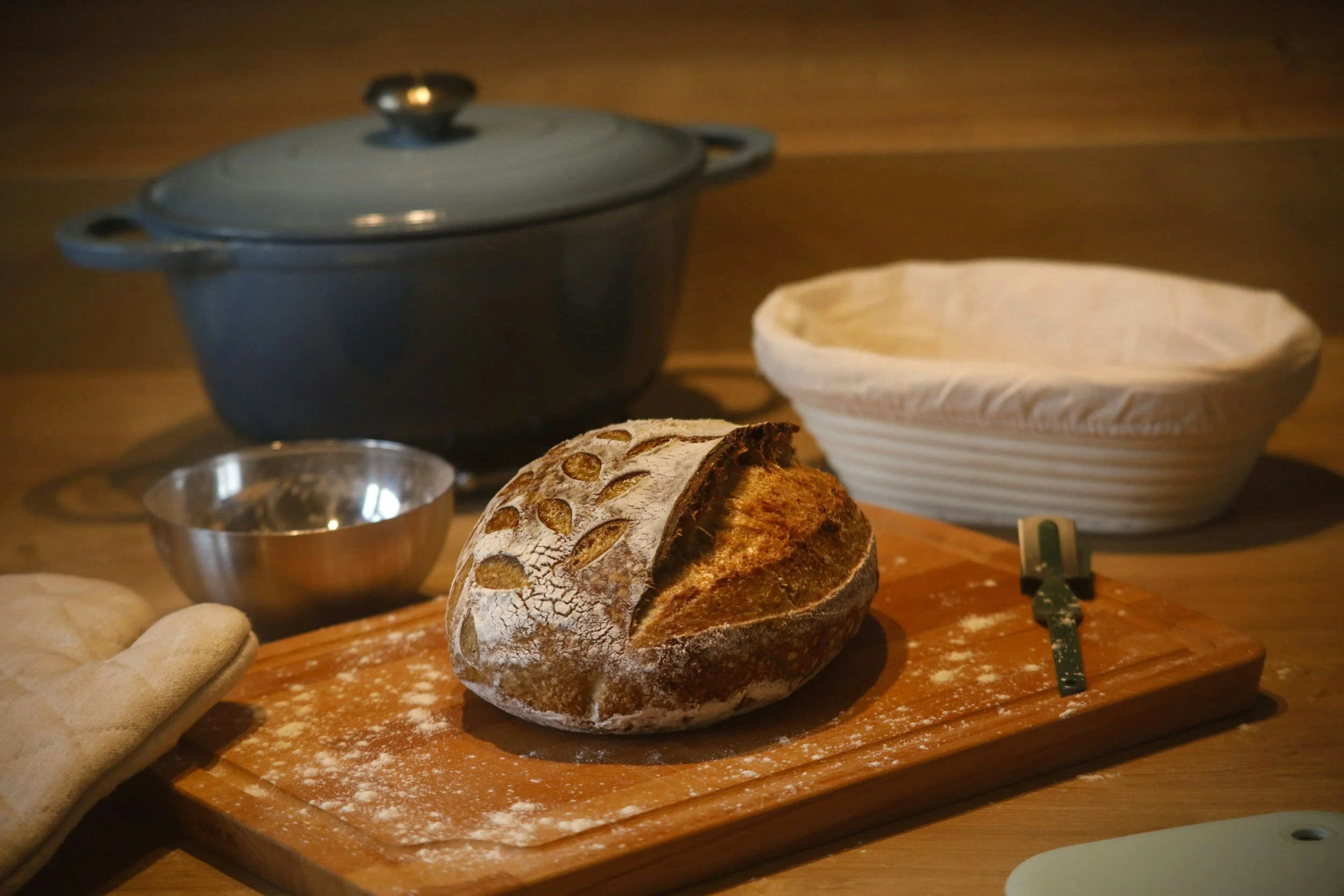 A loaf of rustic bread on a wooden cutting board with flour dusted on top and a crusty exterior. Behind the bread, there is a small metal bowl, a banneton proofing basket, and a cloth. In the background, a large cast iron Dutch oven with a lid and a handle is visible on a wooden surface, with a wooden countertop or table behind it.