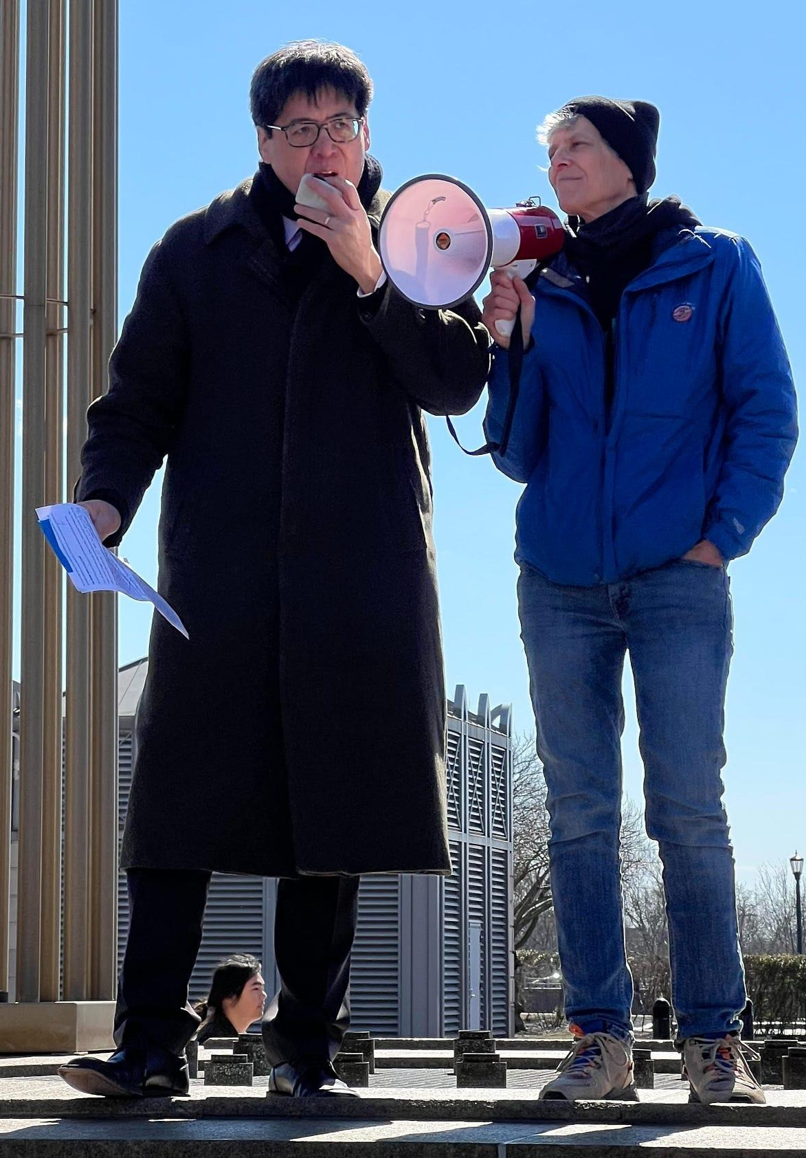 Two individuals standing outdoors, one speaking into a megaphone and holding a piece of paper, the other listening with his hands in his pockets, under a clear blue sky.