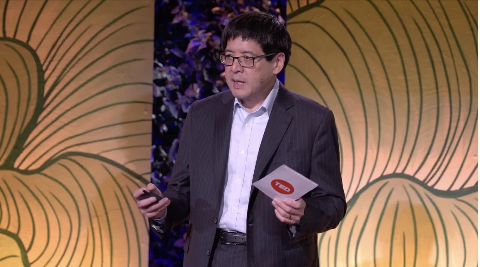 Sam Wang giving a TED Talk on stage, wearing a suit and gesturing while holding a TED speaker card.