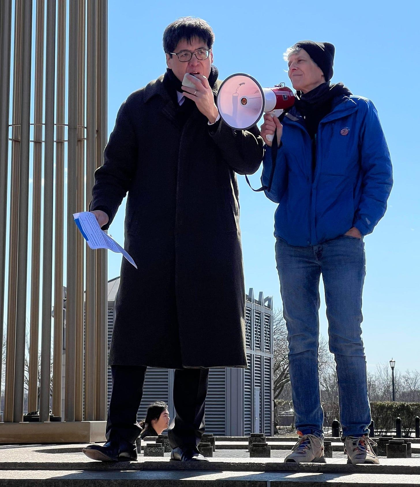Two individuals standing outdoors, one speaking into a megaphone while holding papers, the other listening, with a clear blue sky in the background.