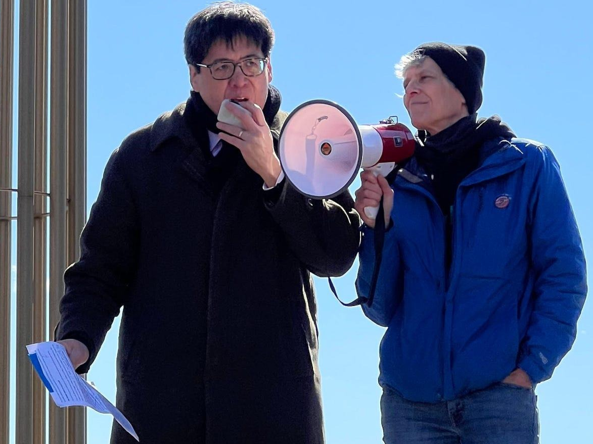 Sam Wang speaks into a handheld microphone while standing beside another person holding a megaphone at an outdoor rally.