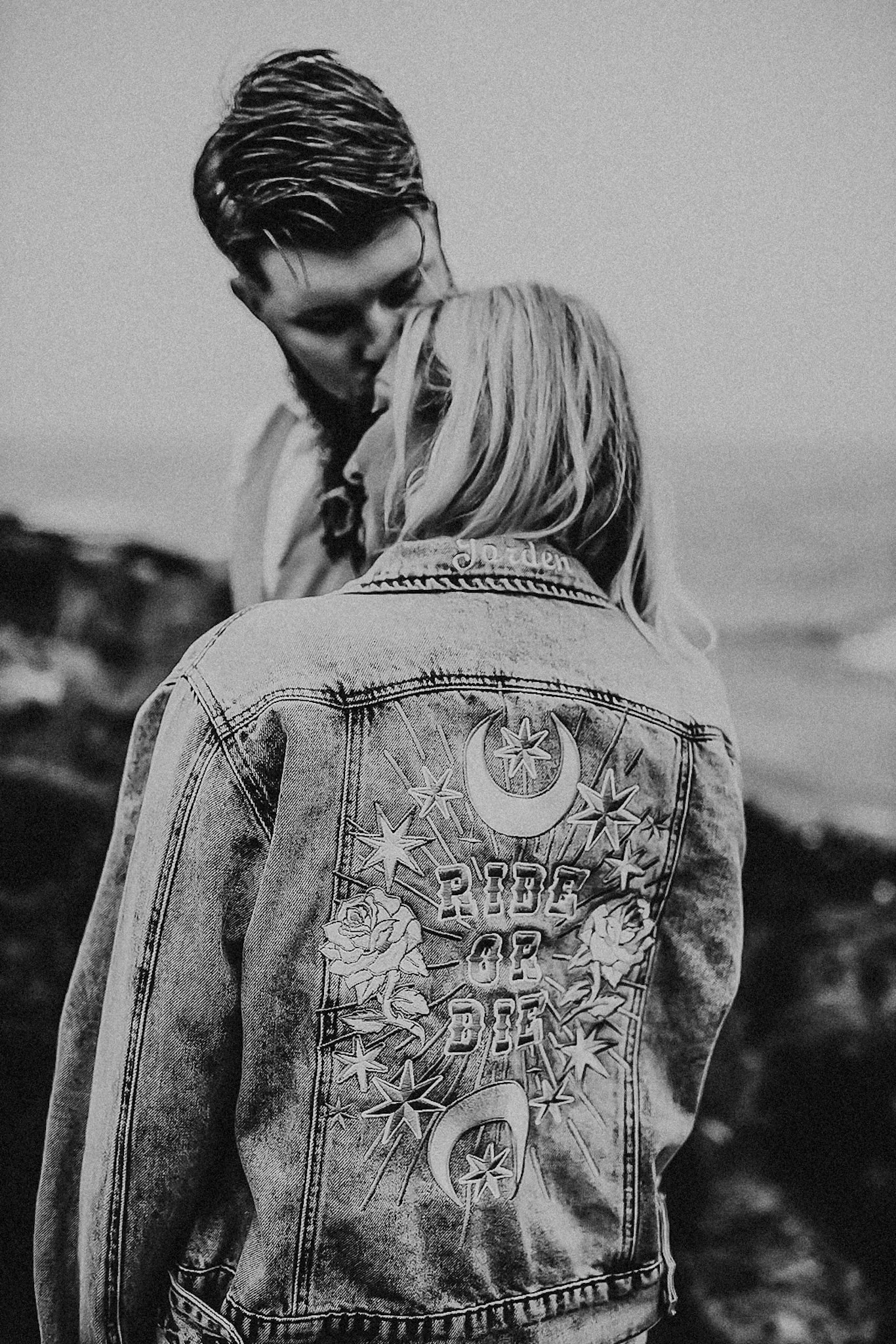 A black and white photo of two women, one with short, dark hair and the other with long blonde hair, standing close together outdoors. The woman with long blonde hair is seen from the back, wearing a denim jacket with a large embroidered design on th