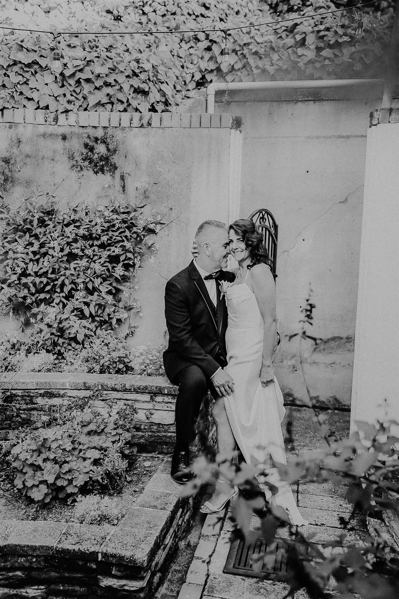 A bride and groom in wedding attire sharing a moment together in an outdoor setting, with plants, brickwork, and a wall in the background.