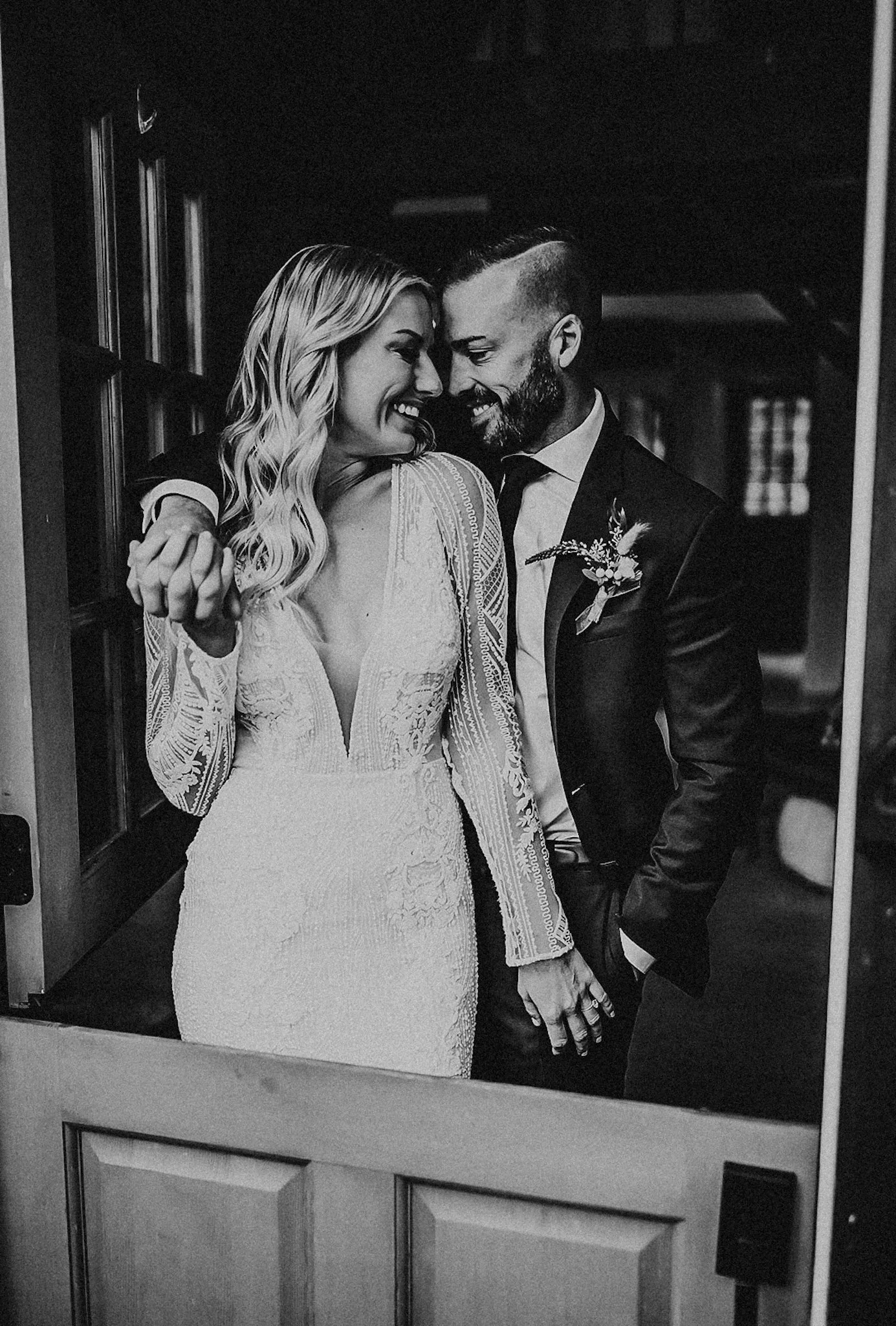 Black and white photograph of a smiling bride and groom in a close embrace, touching foreheads and holding hands, inside a building near a door.