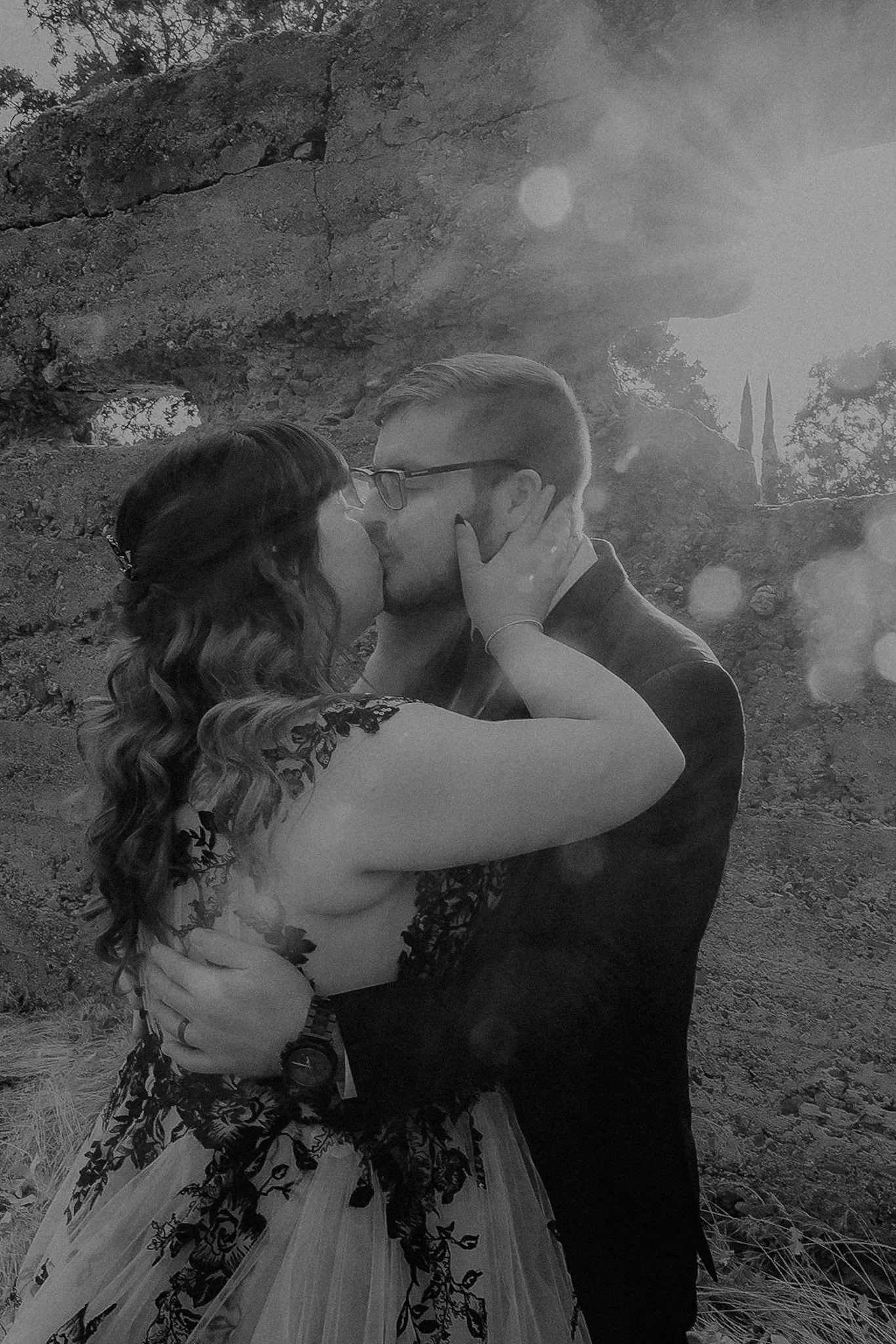 A couple kissing outdoors, surrounded by rocks and trees, with sunlight shining through.