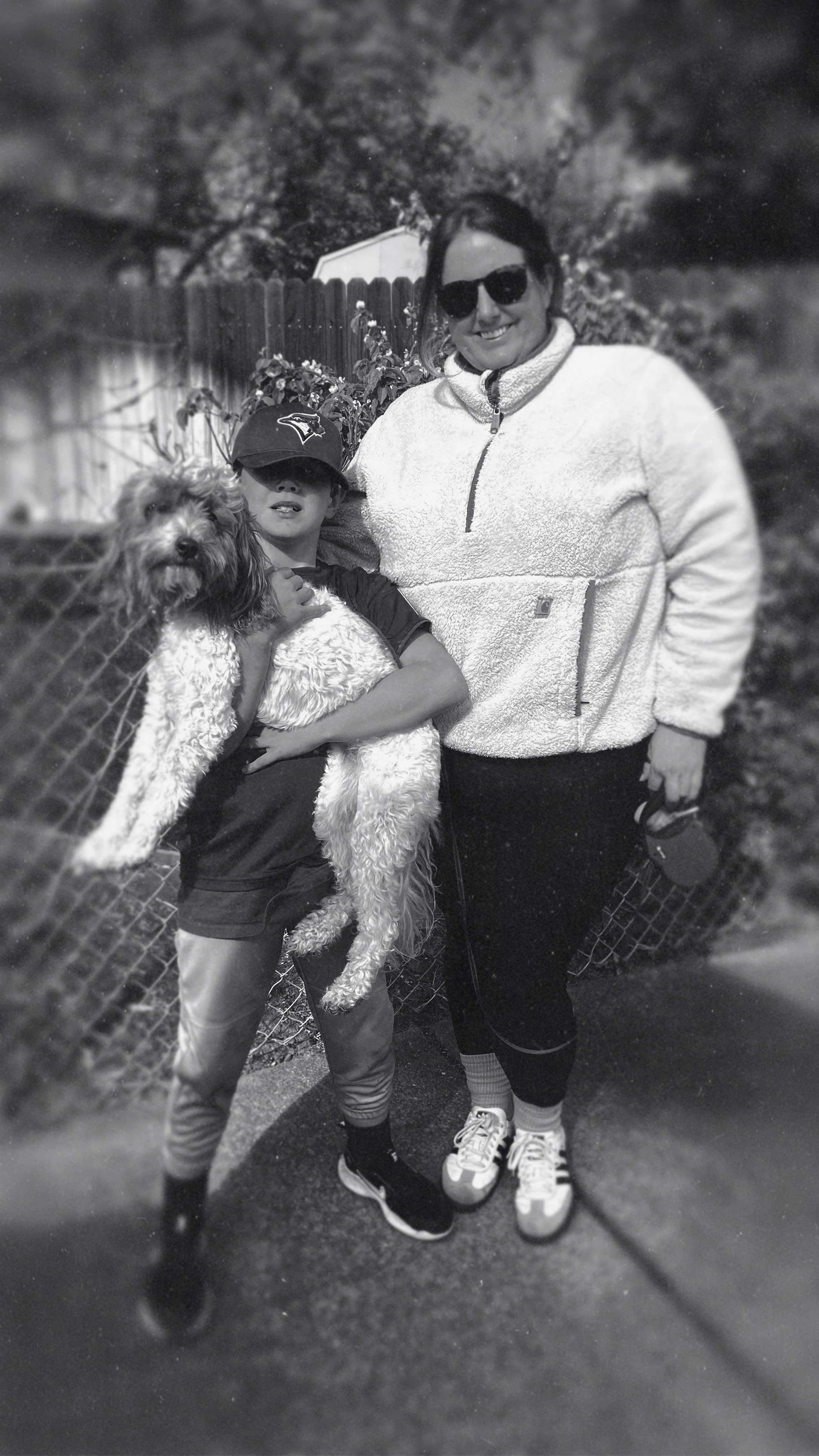 A woman and boy pose outdoors for a photo, with the boy holding a dog. The woman wears sunglasses and a fleece jacket, while the boy wears a baseball cap and casual clothing. They are standing on a sidewalk with a fence and trees in the background.