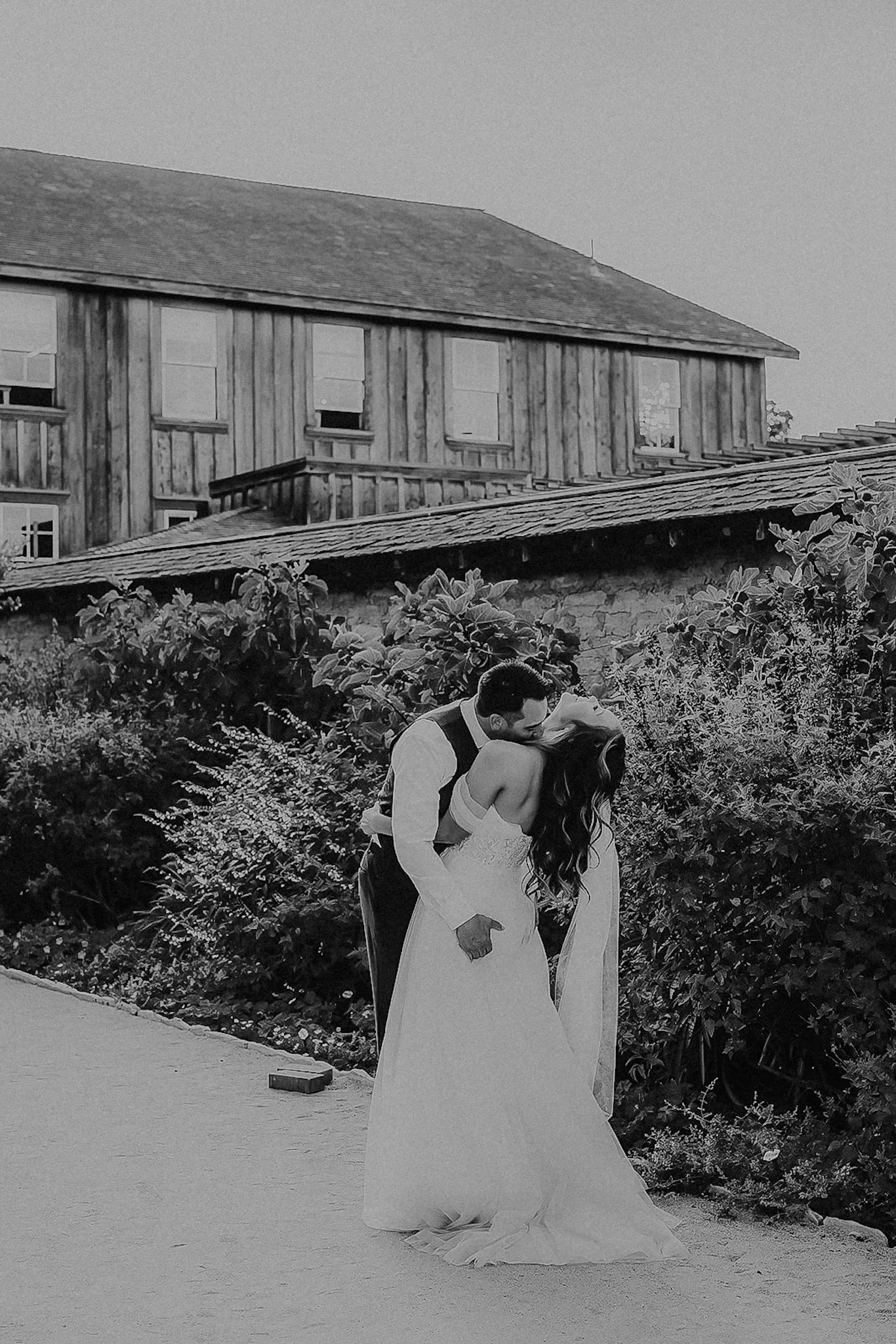 A black and white photo of a newlywed couple kissing outdoors. The groom is wearing a vest and shirt, and the bride is in a strapless wedding gown with long hair. They are standing near some bushes, with a rustic building in the background.