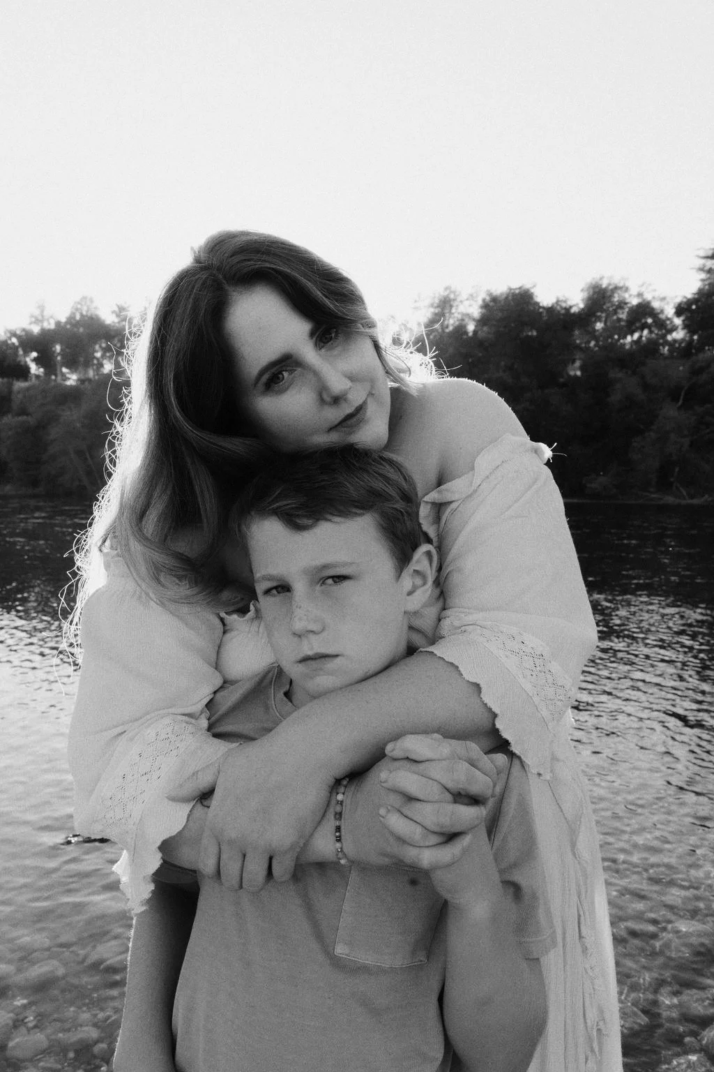 A woman and boy are embracing near a body of water, with trees in the background. The photo is in black and white.